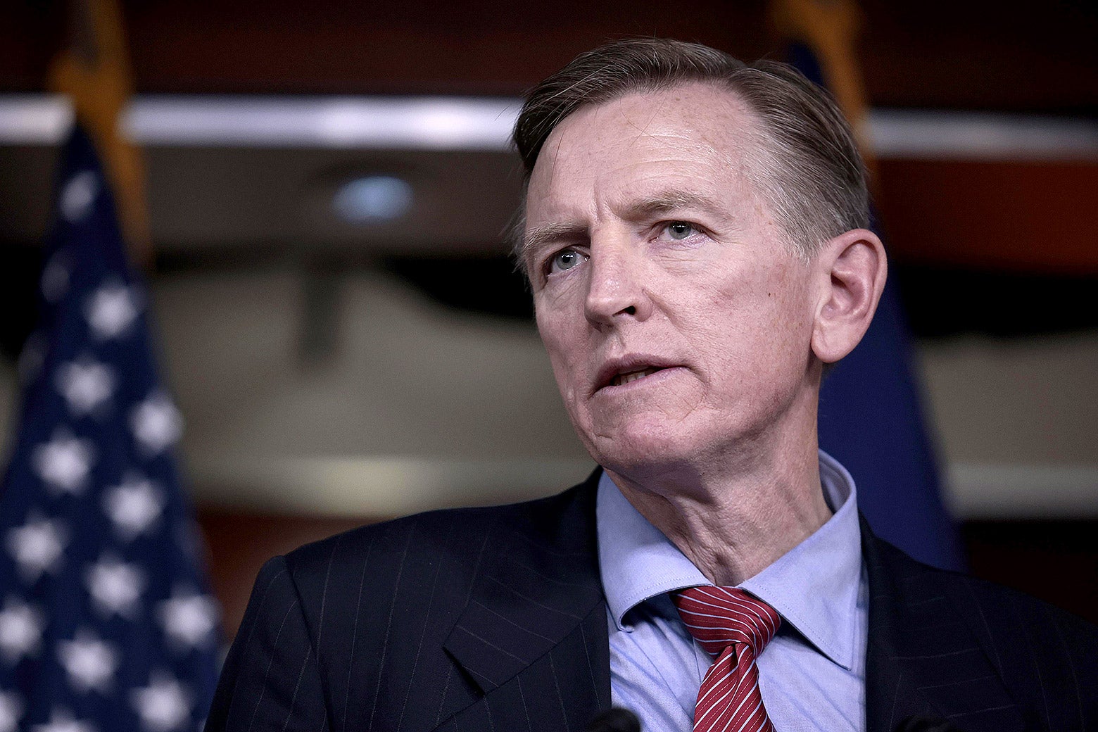 Gosar, wearing a suit jacket, a blue shirt, and a red-striped tie, speaks in front of an out-of-focus American flag.