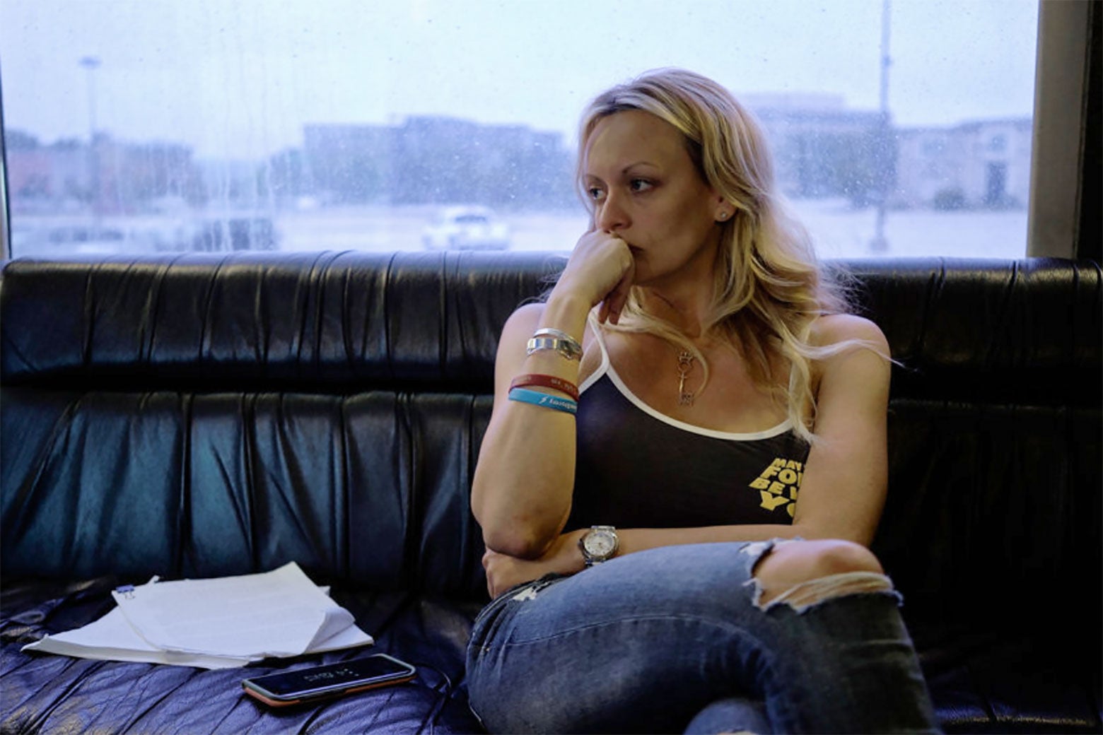 Stormy Daniels sits on a couch and looks to the side, hand to her face in concern. There is a cellphone and papers on the couch next to her.