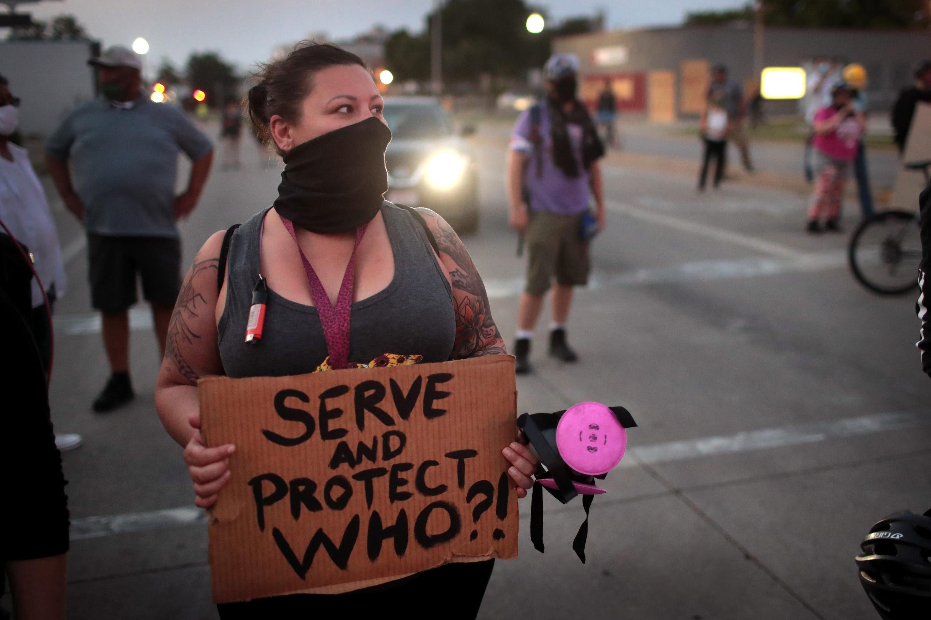 A masked woman holding a sign that reads "Serve and Protect Who?!" stands among other protesters in the street