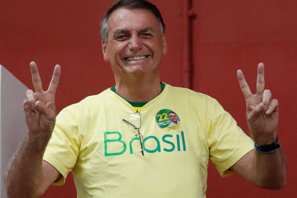 Bolsonaro, wearing a yellow t-shirt in front of a red backdrop, flashes peace signs with both hands.