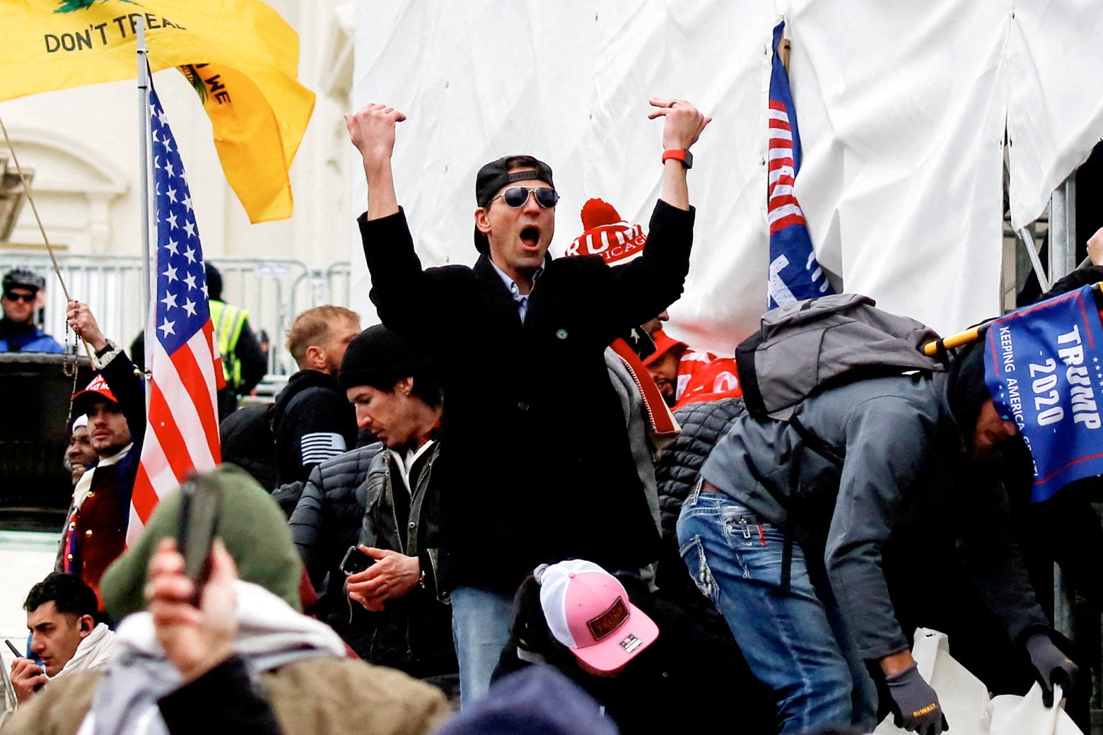 Kelley, wearing black sunglasses and a backward hat, makes a "come this way" gesture with two hands against a backdrop of other Jan. 6 rioters and white sheets over scaffolding.