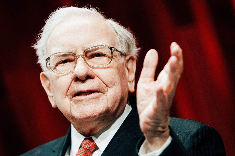 Warren Buffett, wearing a suit, raises his hand.