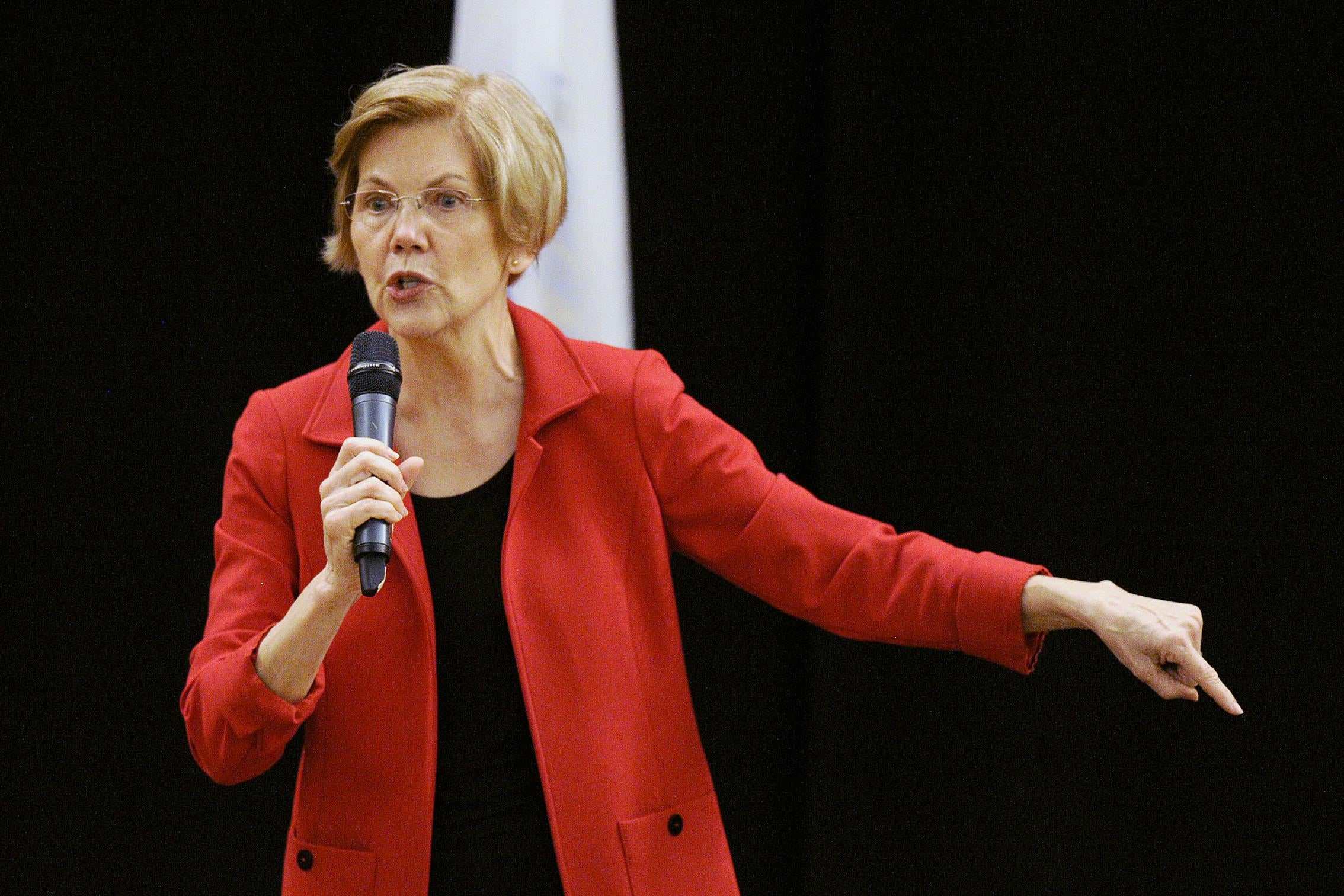 Sen. Warren, dressed in a red jacket, holds a microphone in one hand and with the other gestures toward the ground.