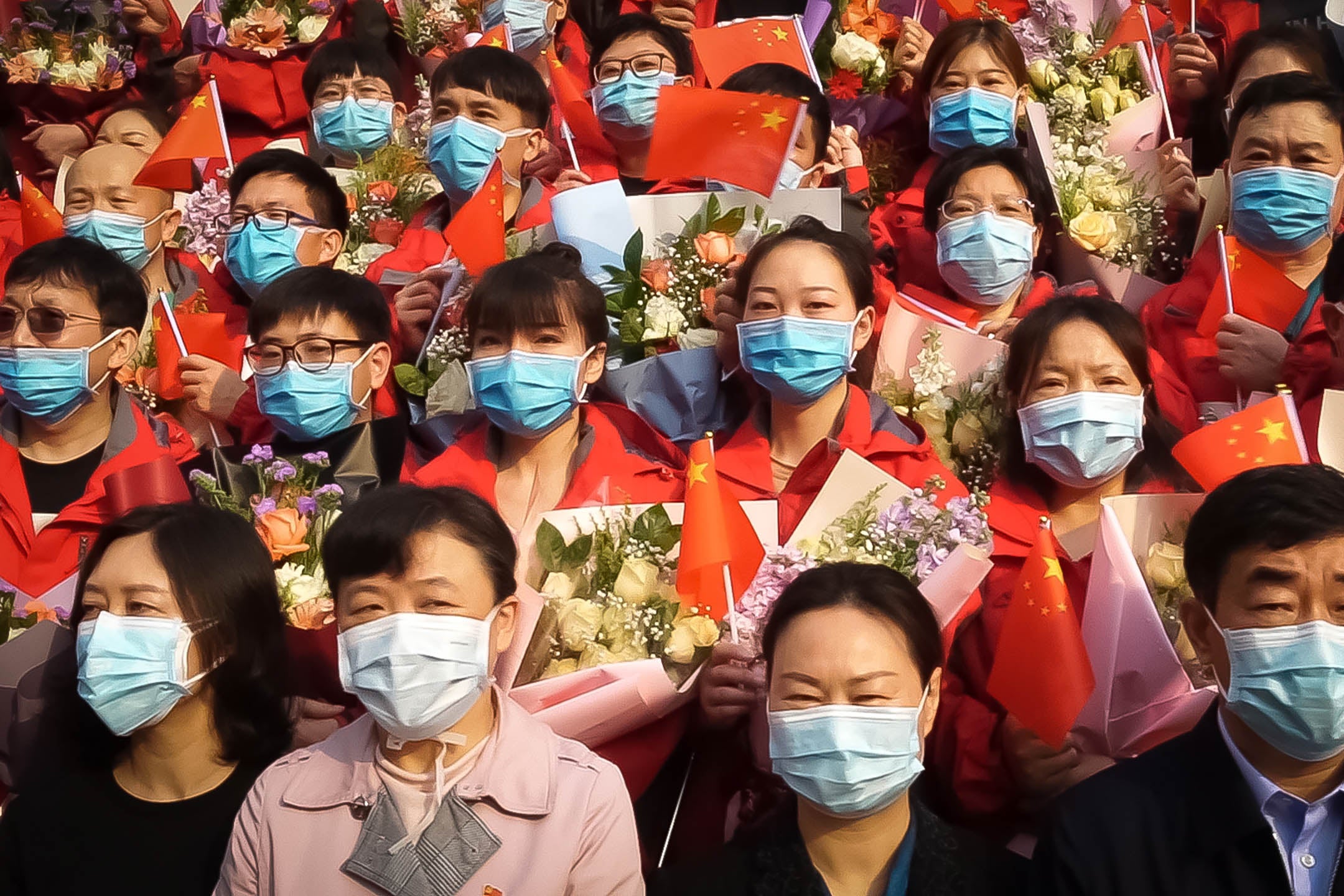 A crowd of Chinese people all wearing surgical masks and holding up little Chinese flags