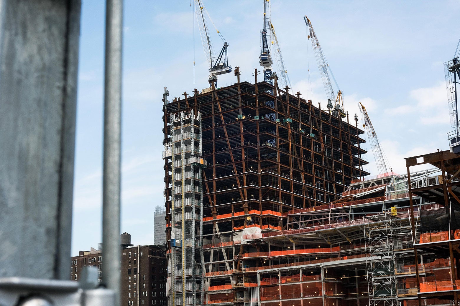 A construction site for what appears to be a new apartment building. The tower being constructed is flanked by cranes, and bright orange construction equipment is visible on the lower levels.