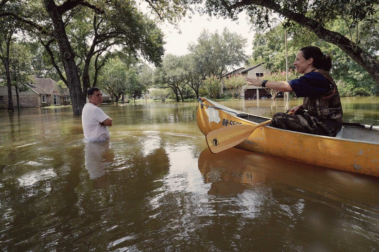 A woman in a canoe talks to a man standing in hurricane floodwaters. Houses and trees are partially submerged in the background.