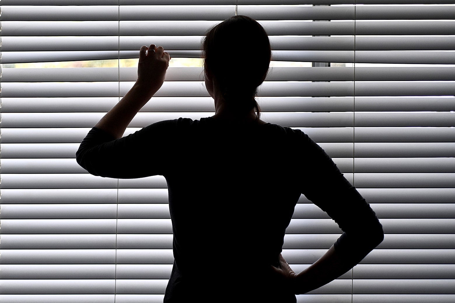 A silhouette of a woman in a dark room peeping through shutters at glimpses of sunlight