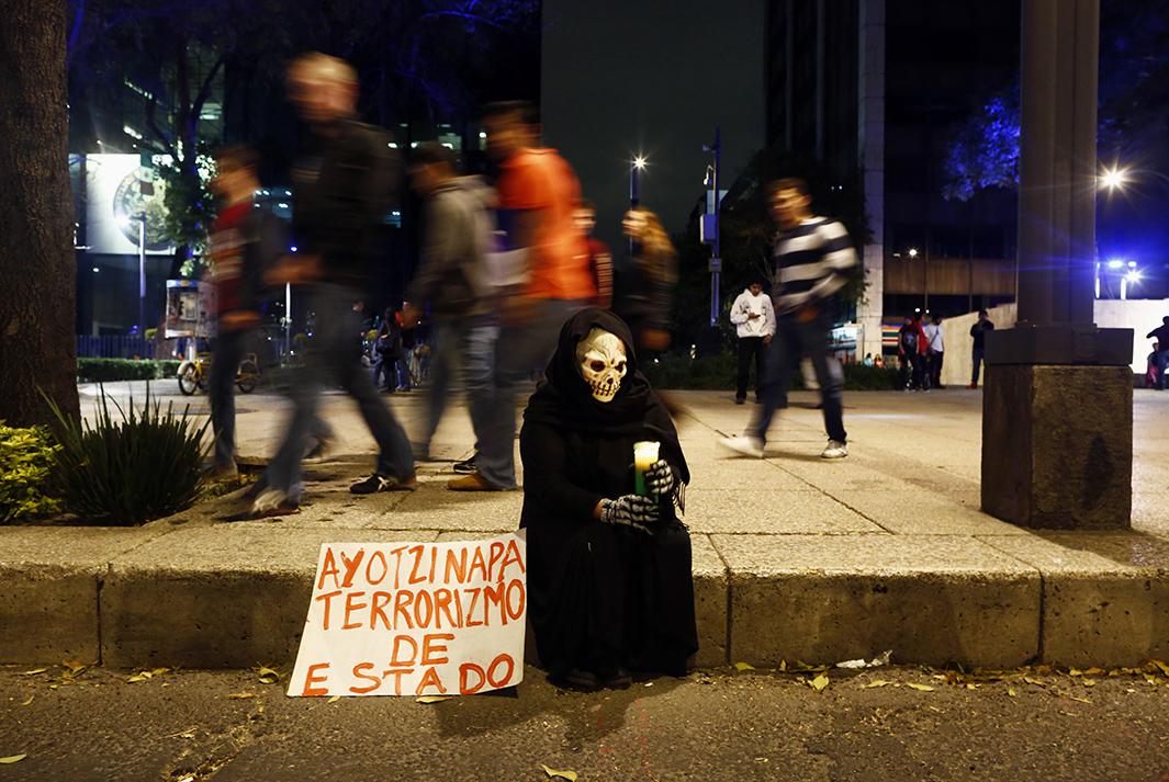 A masked protester sits on a sidewalk holding a candle after the Nov. 8 protest. The sign reads "Ayotzinapa, state terrorism". 