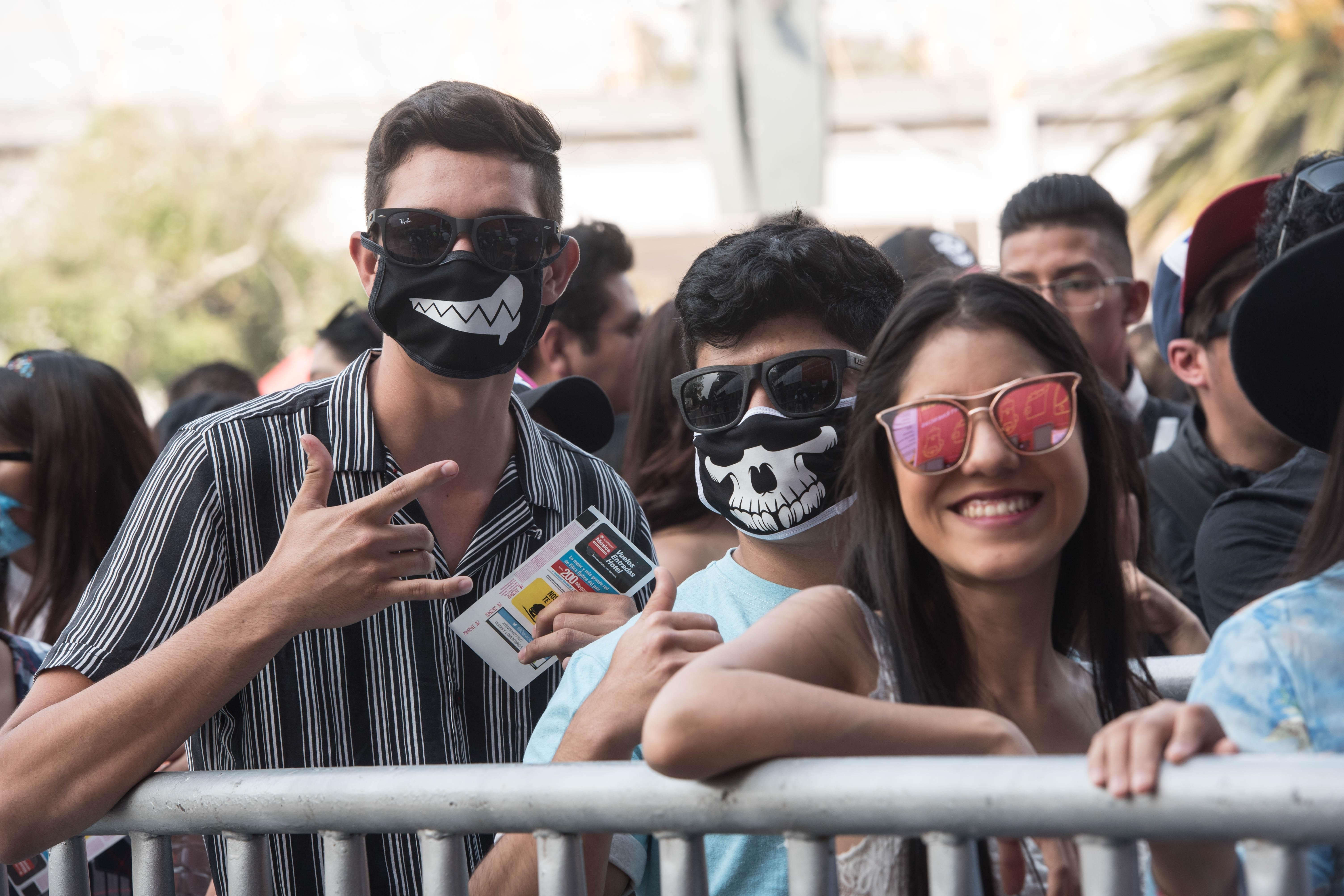 A crowd of concertgoers, some wearing face masks with skull imagery, outside on a sunny day