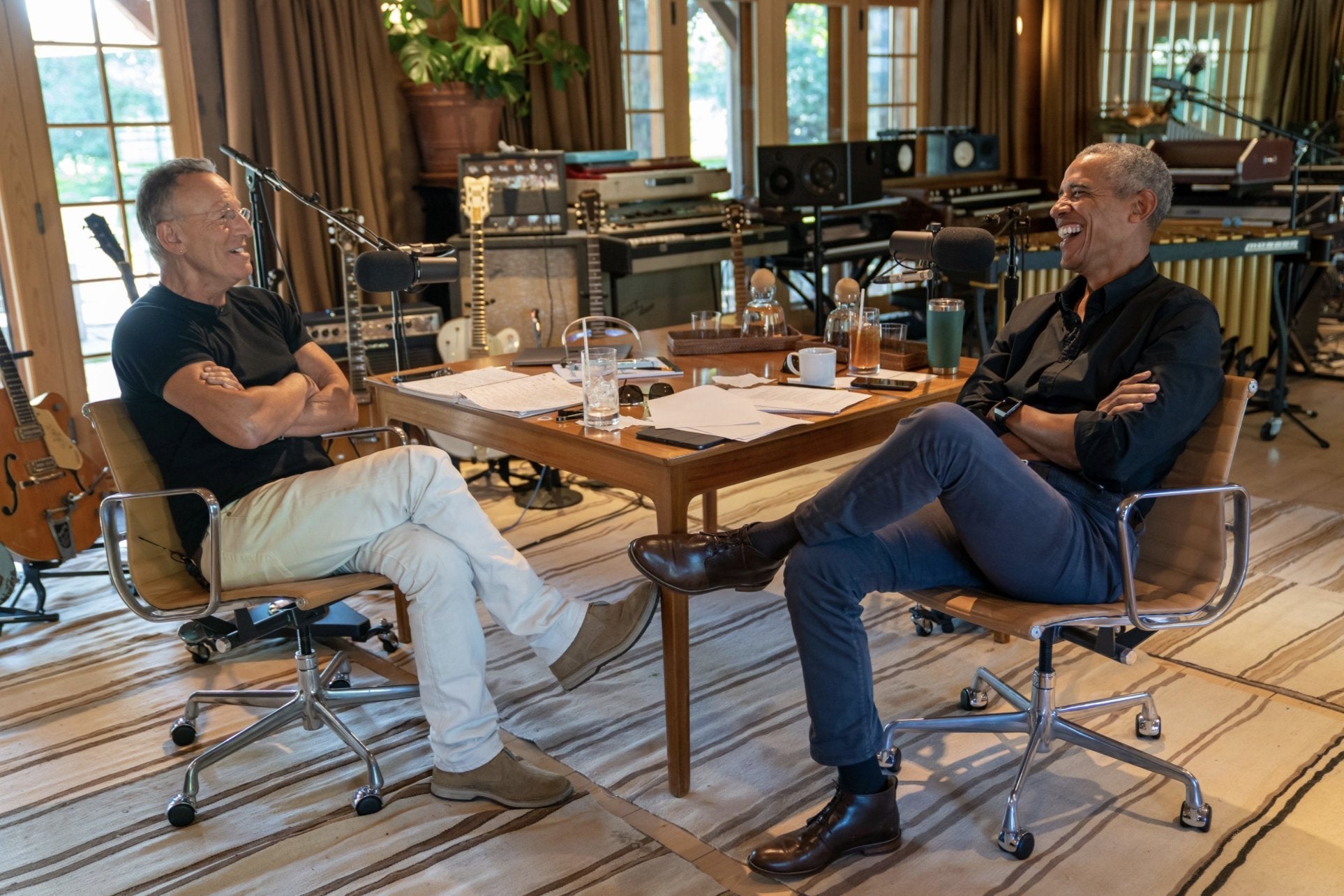 Bruce Springsteen and Barack Obama sit facing each other at a table covered in many beverages and papers. They are smiling into recording microphones. There are many musical instruments behind them.
