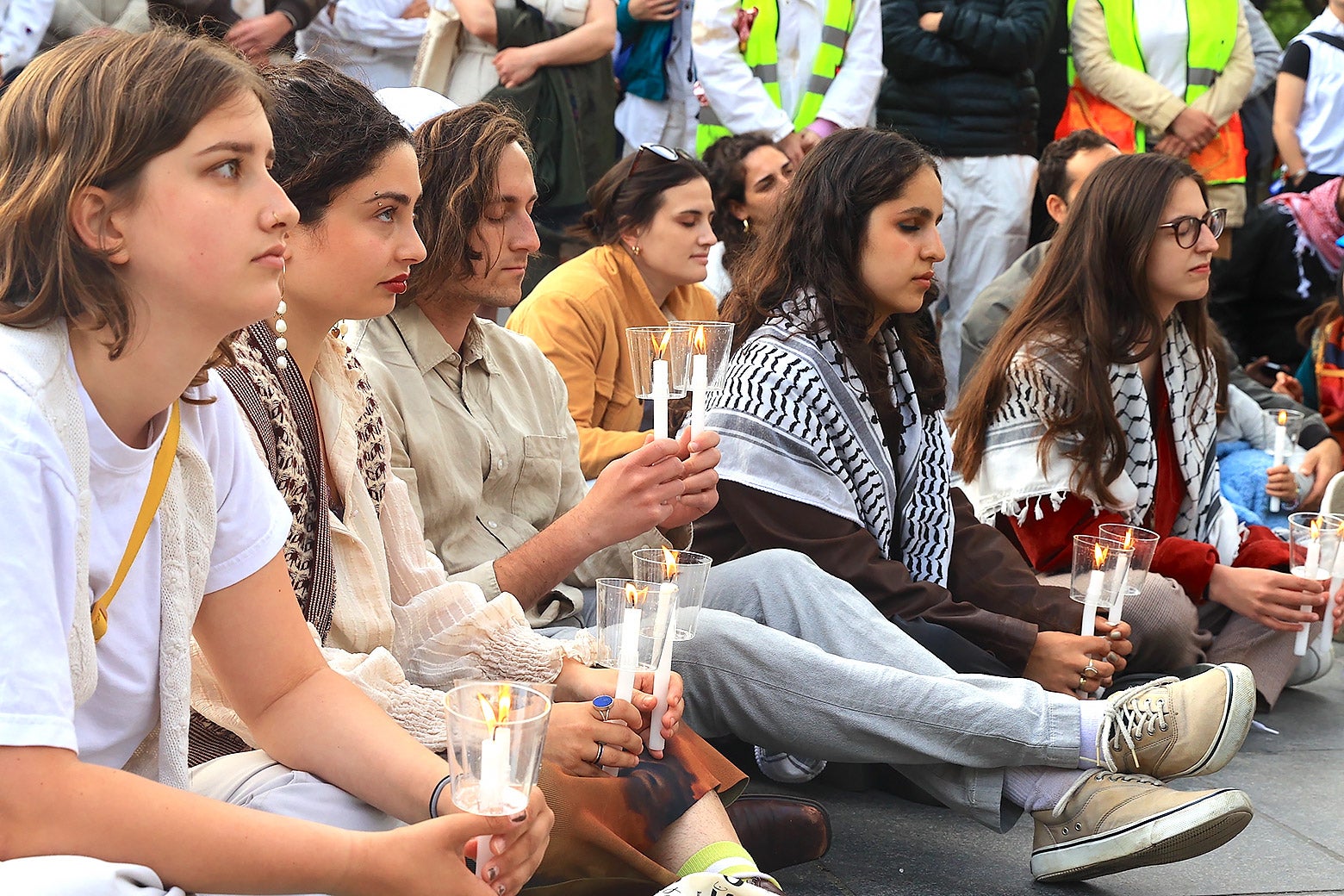 Seated Jews, young men and women, hold Shabbat candles; two of the women wear kaffiyeh draped over their shoulders.