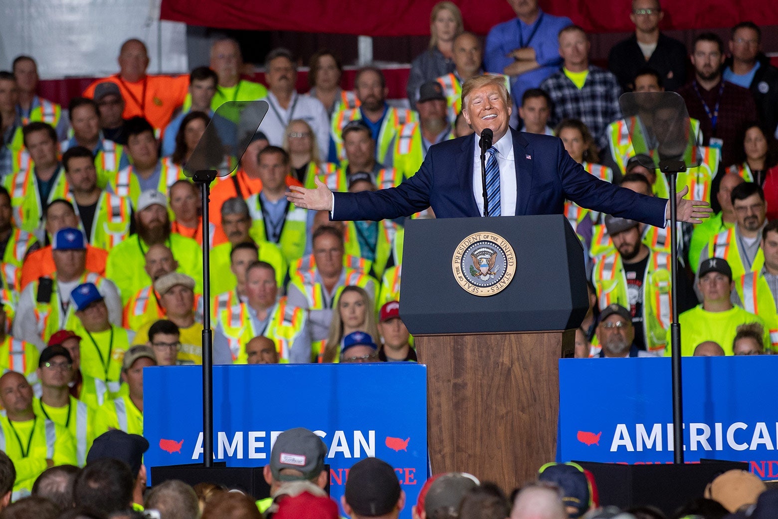 Trump speaking at a podium onstage with an audience of workers behind him.