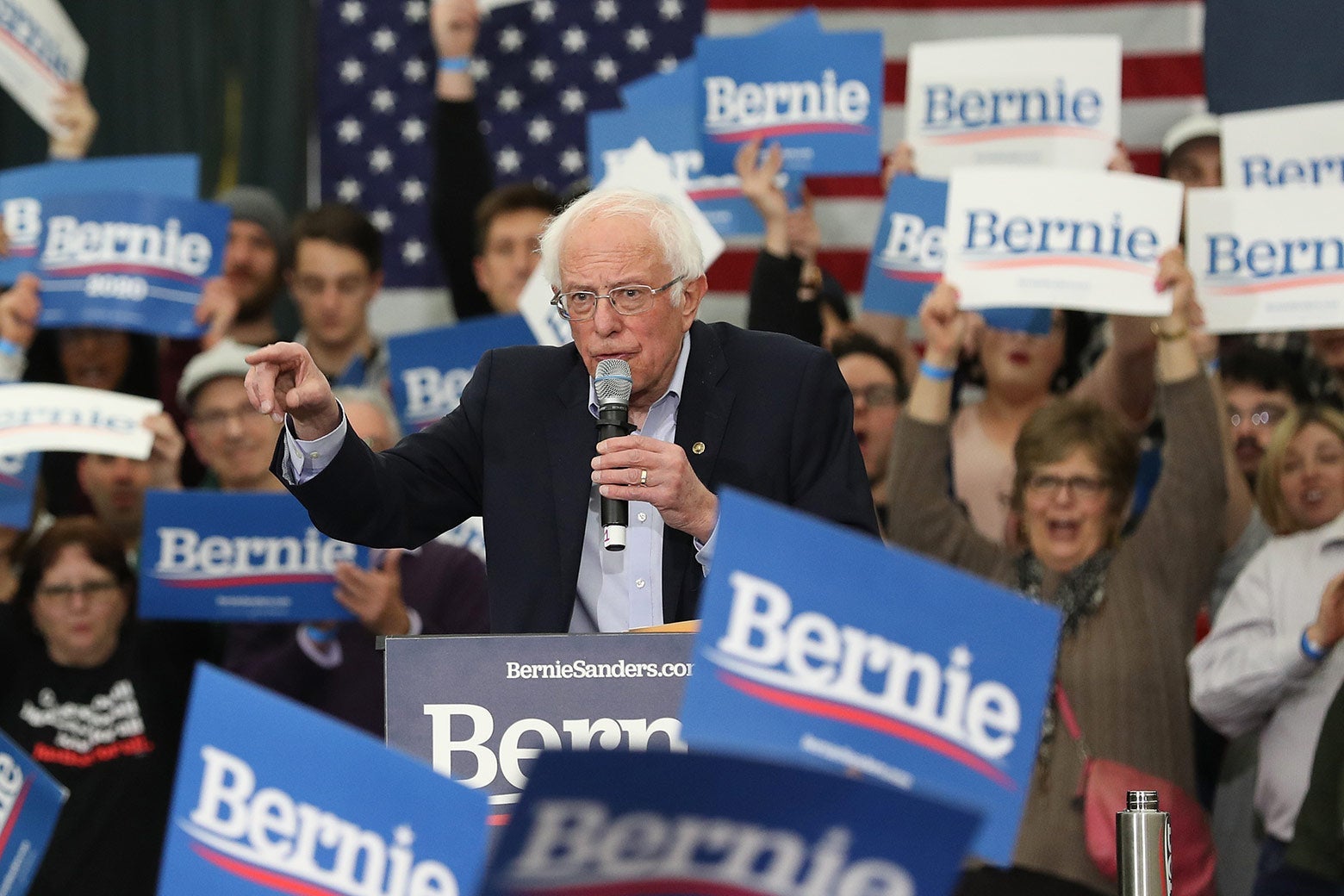 Bernie Sanders at a rally with supporters behind him holding signs that say, "Bernie."