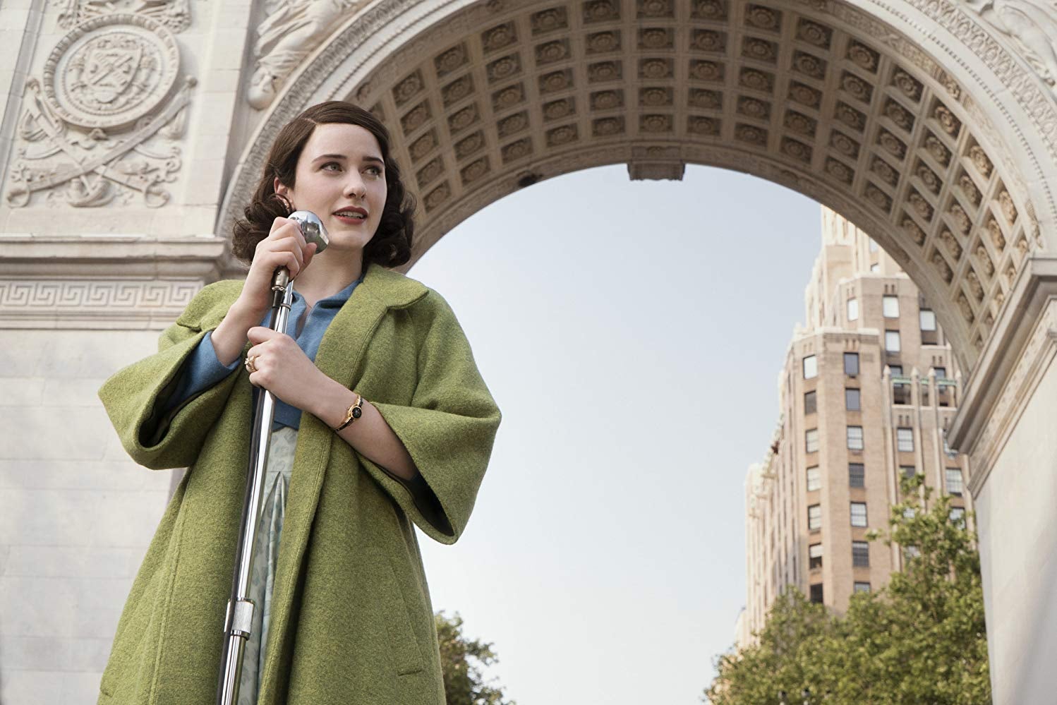 Rachel Brosnahan stands on front of a microphone wearing a green coat.