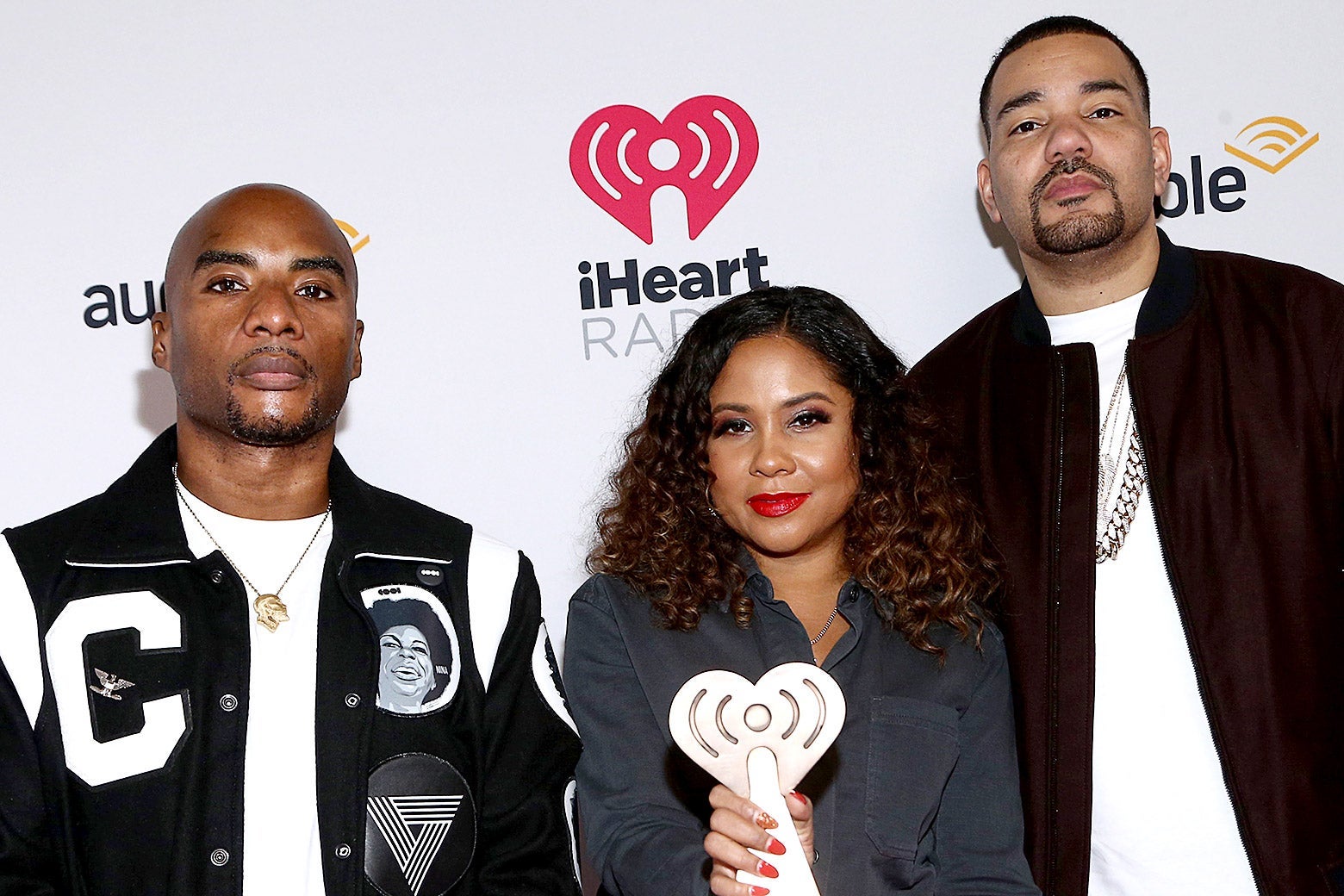 Angela Yee holds up a trophy with Charlamagne and Envy standing on either side of her in front of an Audible iHeartRadio step and repeat