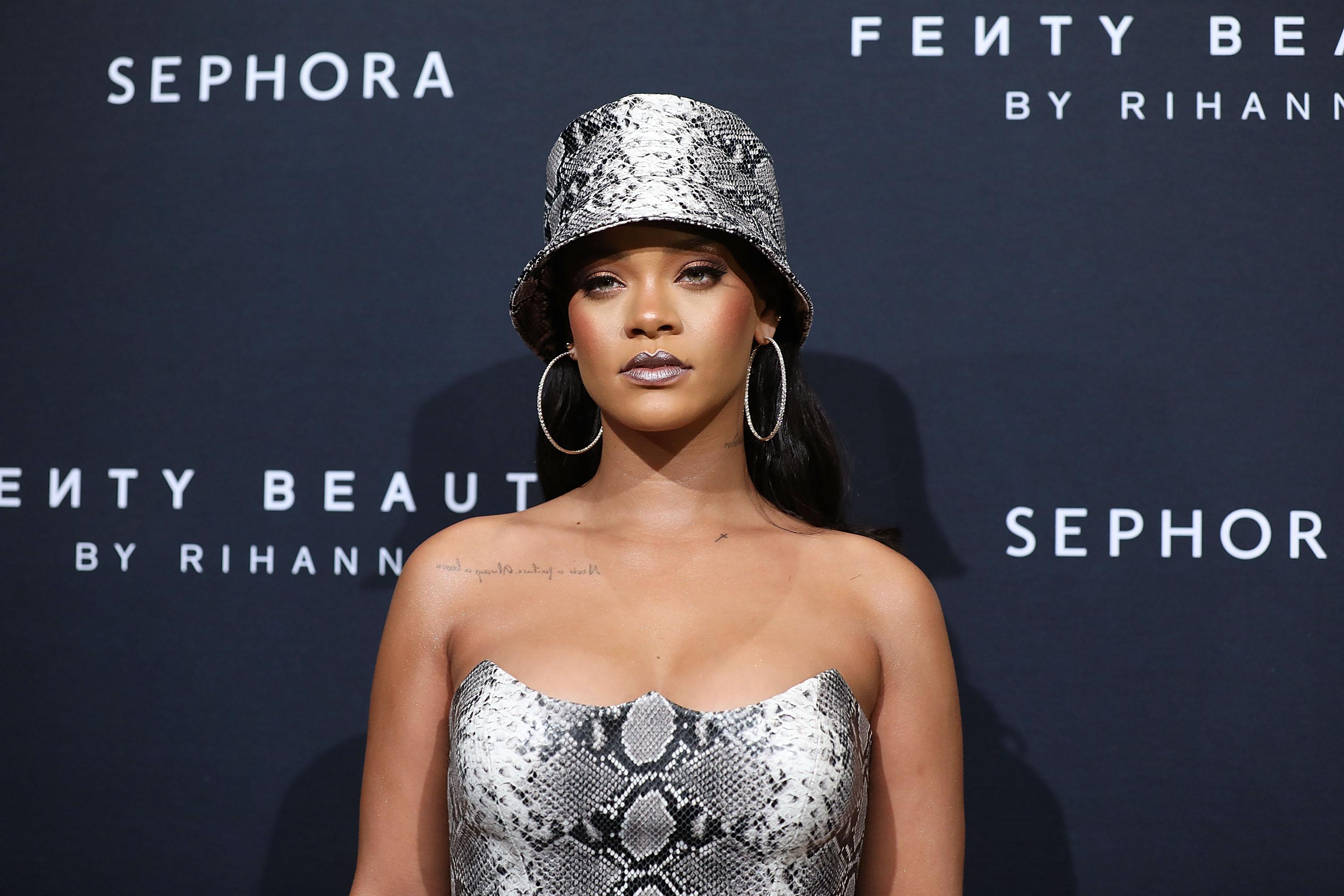 Rihanna stares straight ahead, wearing a bucket hat and large hoop earrings. Behind her are signs for Fenty Beauty and Sephora.