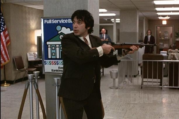 Al Pacino, dressed in a suit, stands holding out a gun in a bank. While two people in the background look on.