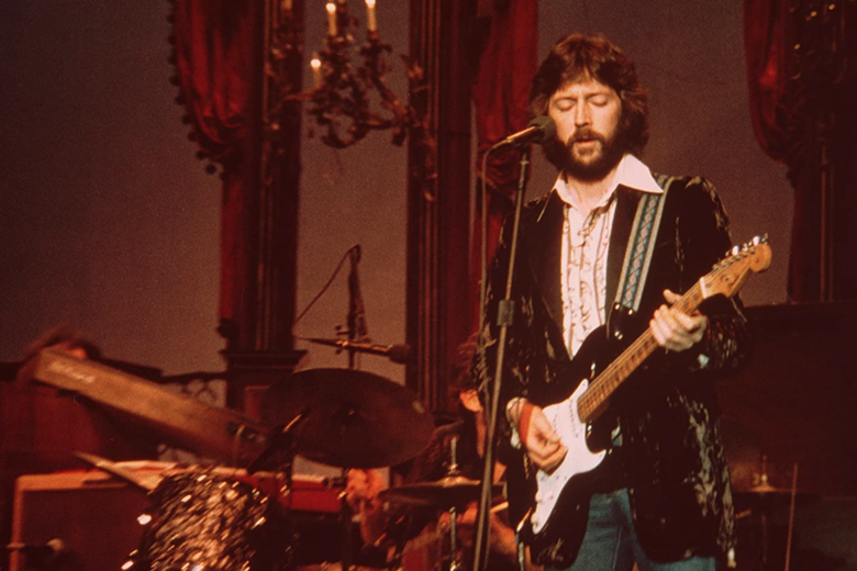 Eric Clapton stands on stage behind a microphone, holding a guitar