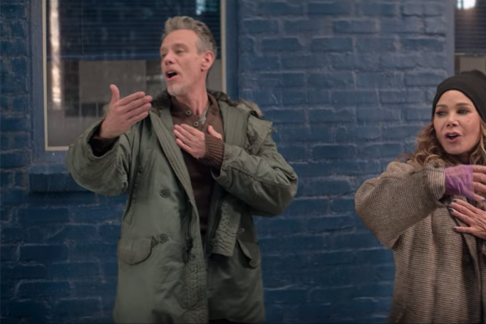Adam Pascal, Daphne Rubin-Vega, and Wilson Jermaine Heredia stand, dressed as homeless people, singing with one arm stretched out and their other hand over their hearts.