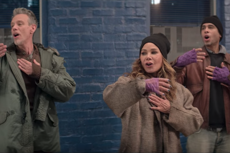 Adam Pascal, Daphne Rubin-Vega, and Wilson Jermaine Heredia stand, dressed as homeless people, singing with one arm stretched out and their other hand over their hearts.