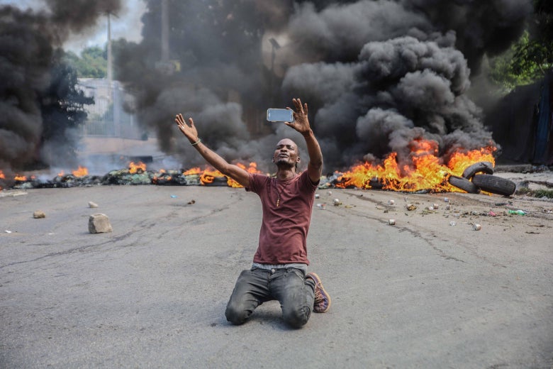 A man kneels in the street with his arms above his head as tires burn behind him.