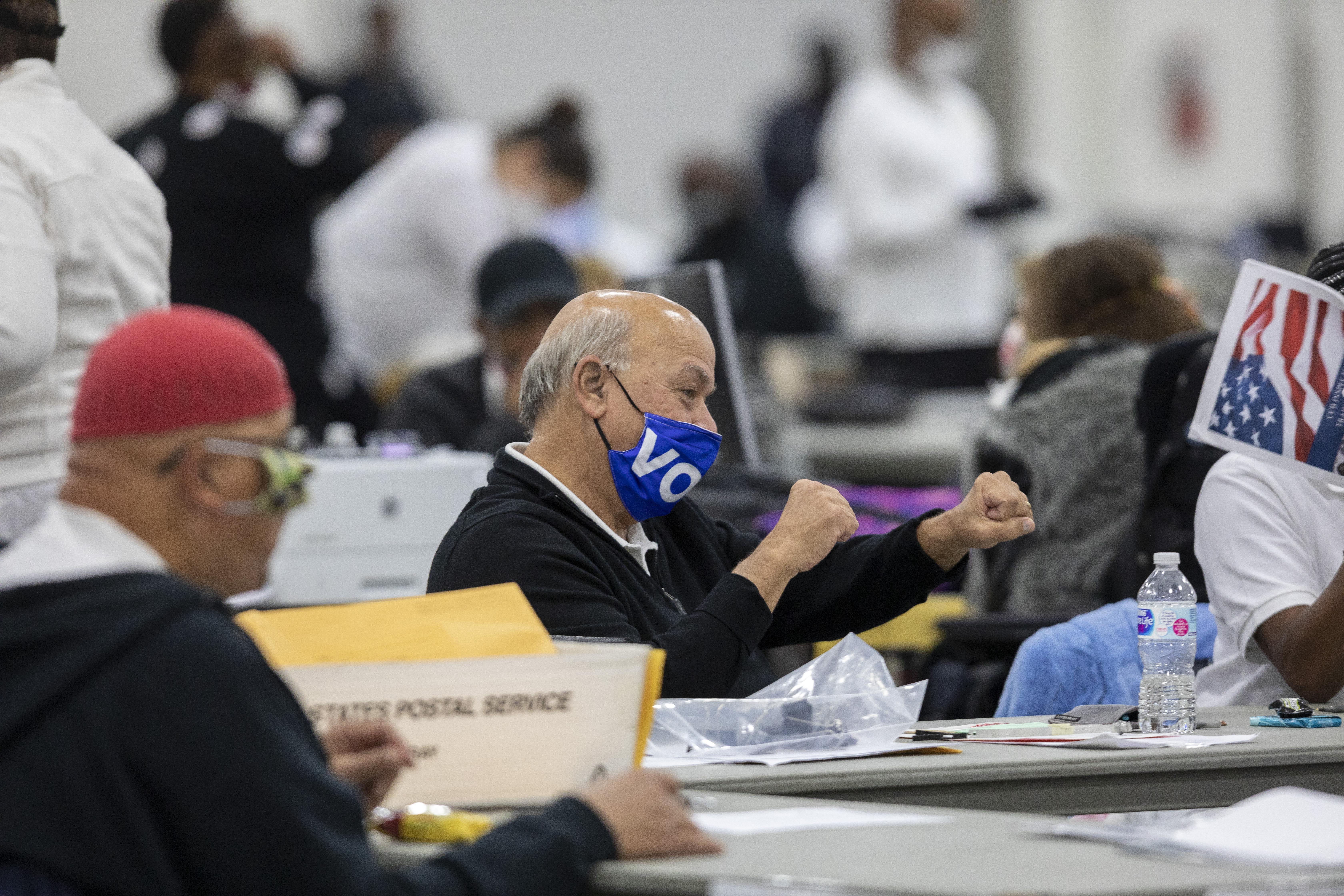 A man wearing a "VOTE" mask pumps his fists in a crowded room of people holding documents and ballots.