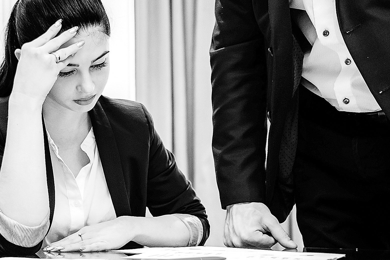 A woman in business attire stares down at some papers on a desk as a man stands over her.