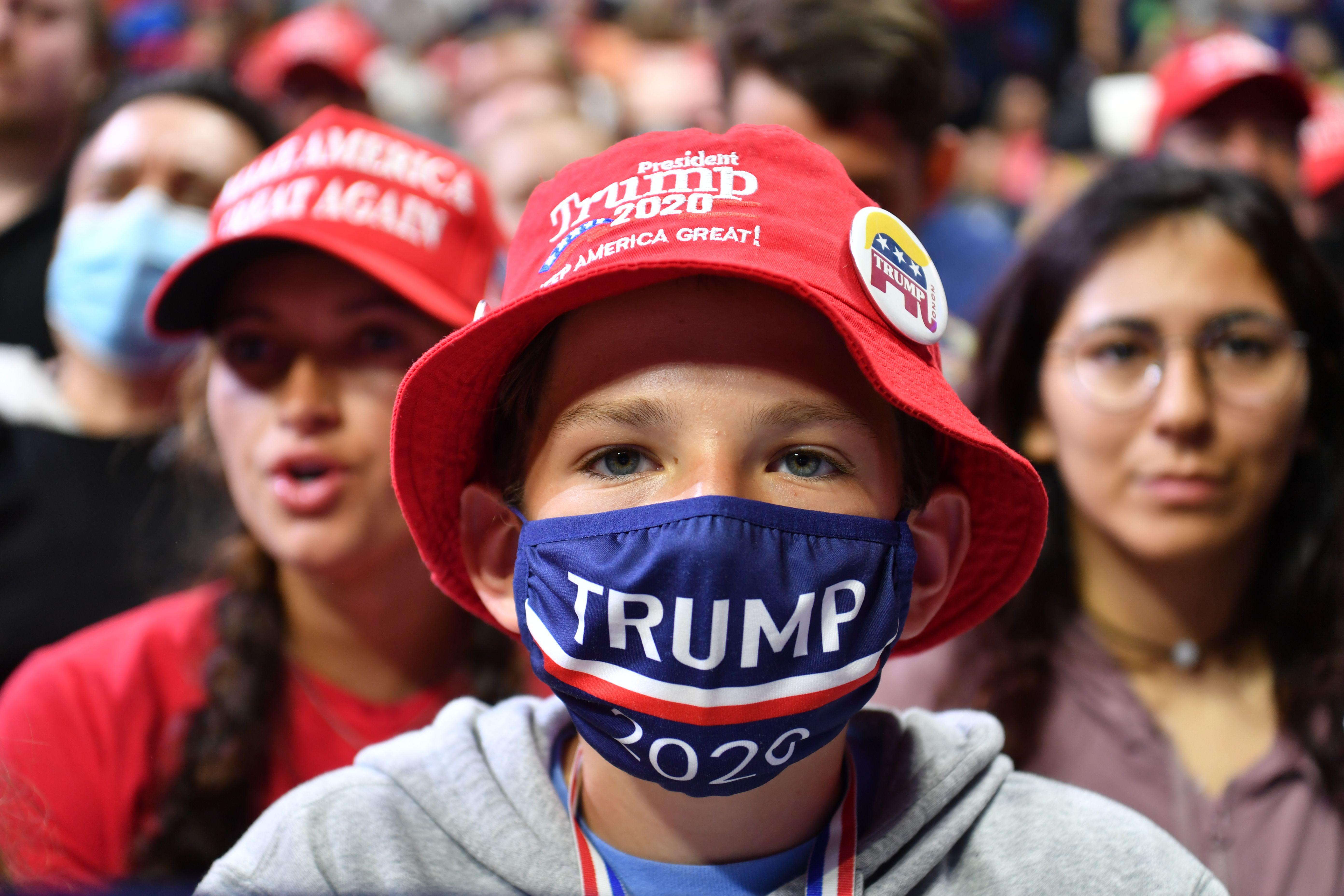 A young boy wears a Trump 2020 mask and a red hat. He's surrounded by others in similar gear.