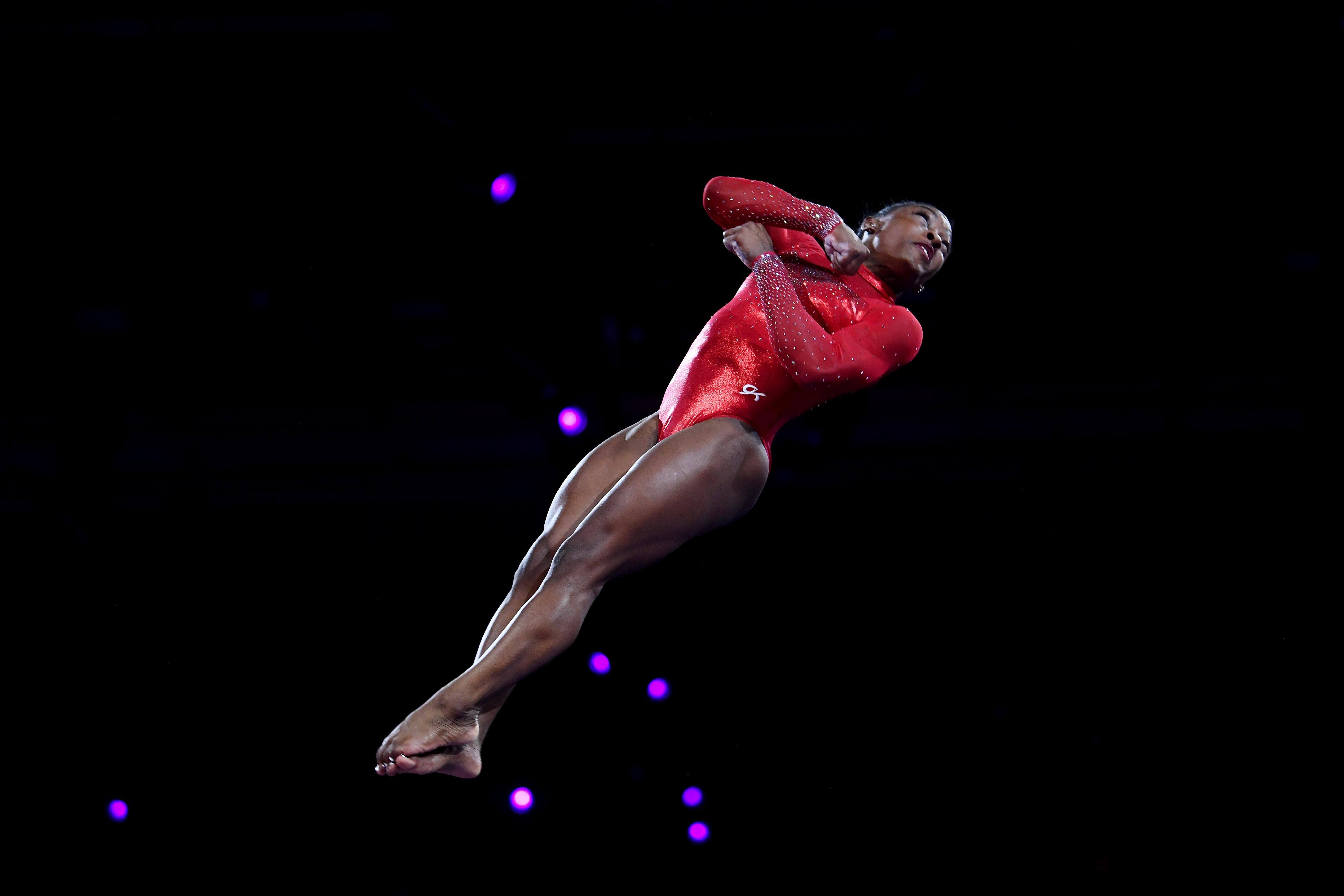 Simone Biles twisting in the air against a black background as she vaults.