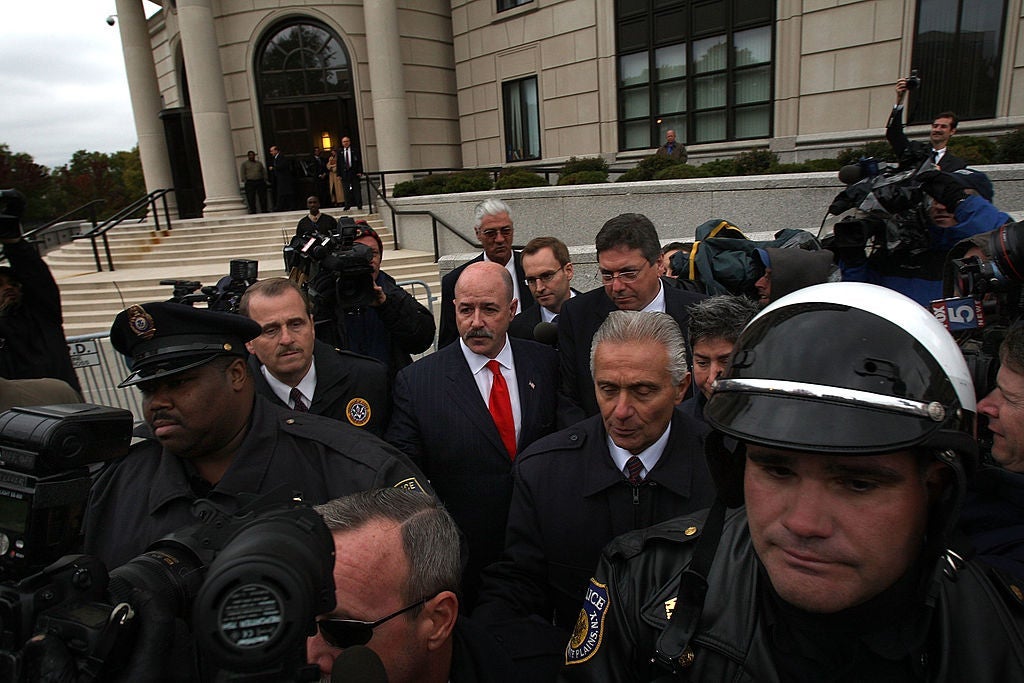 Bernard Kerik, wearing a black suit with a red tie, walks away from a set of courthouse steps while surrounded by police officers and cameras.