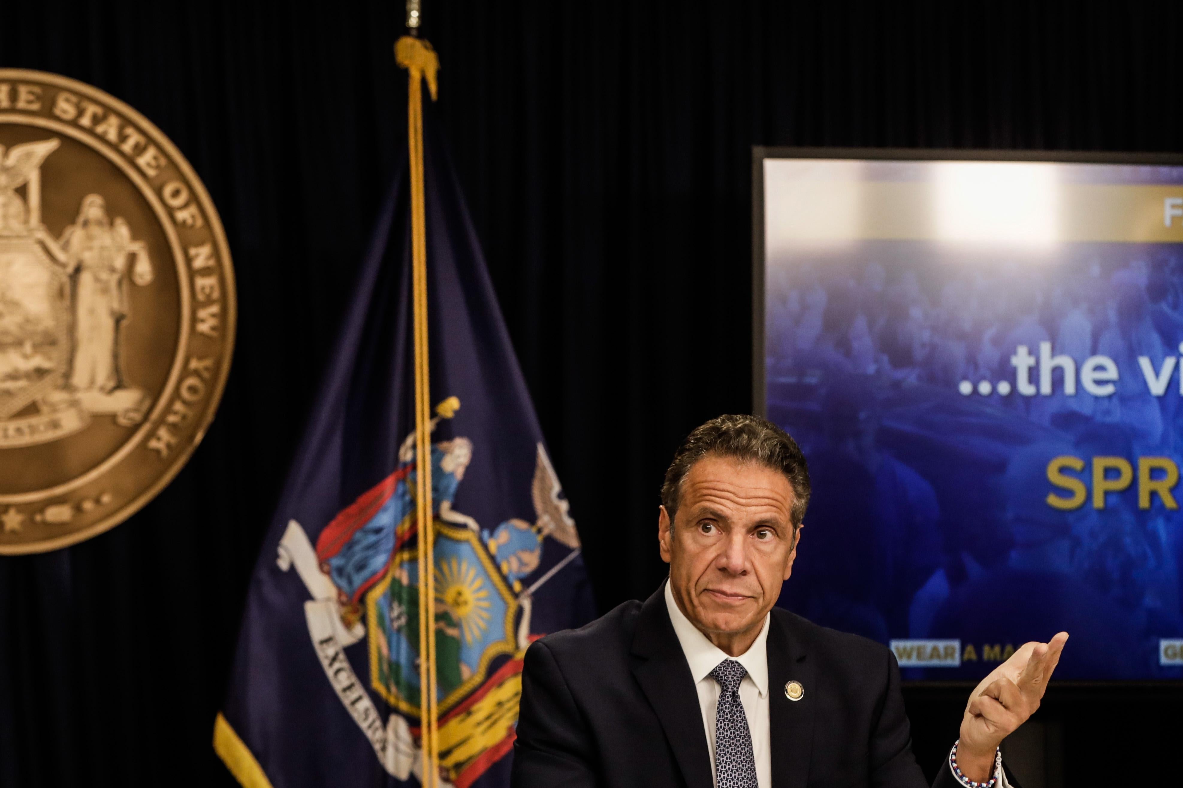 Andrew Cuomo gestures at a screen that says "the virus will SPREAD."