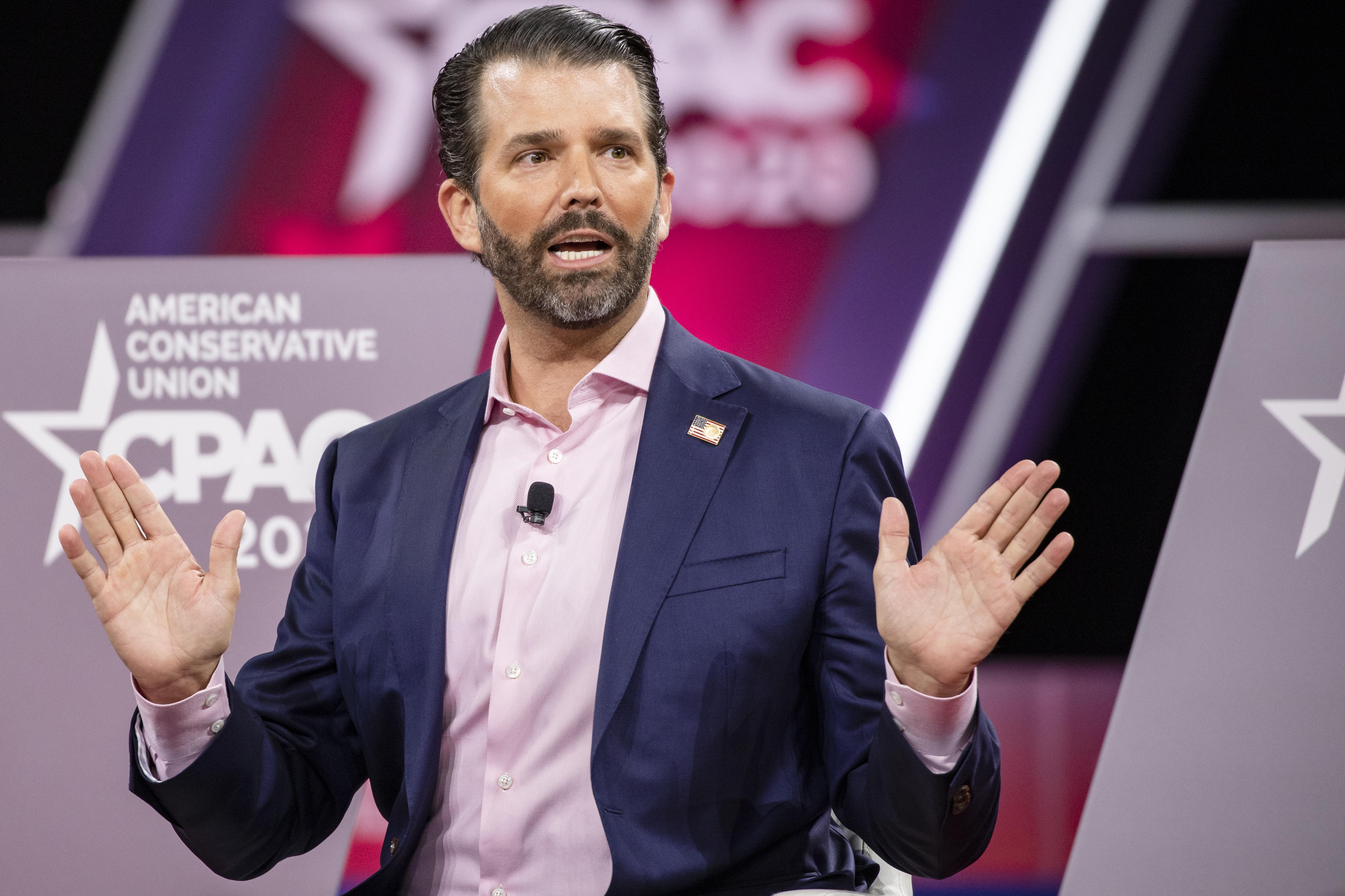 Donald Trump Jr. raises his hands while speaking, standing in front of a wall with a CPAC logo.