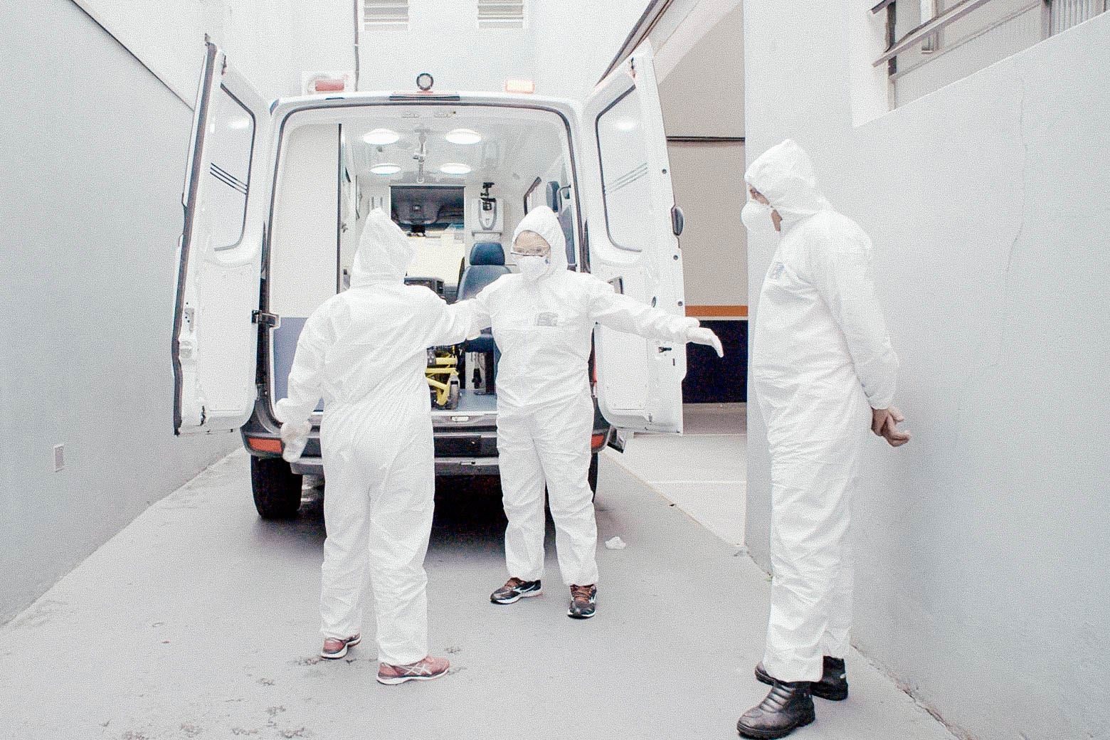Three nurses in white bodysuits stand in front of an open truck trunk in a white corridor.