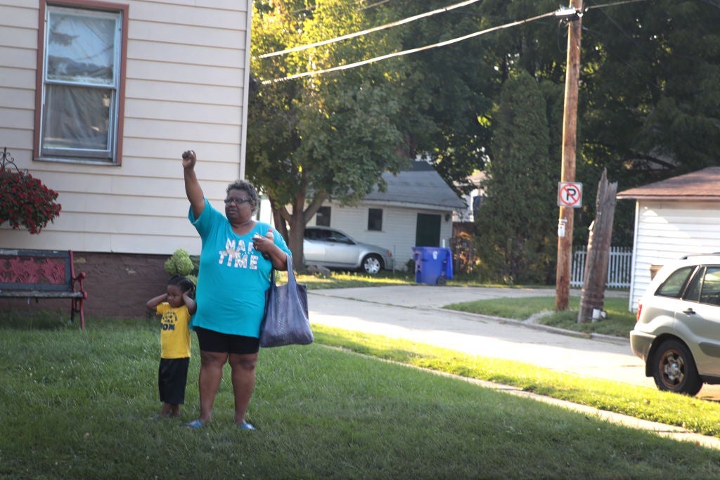 A Black woman standing in the front yard of a house with a toddler holds her right hand up in a fist.