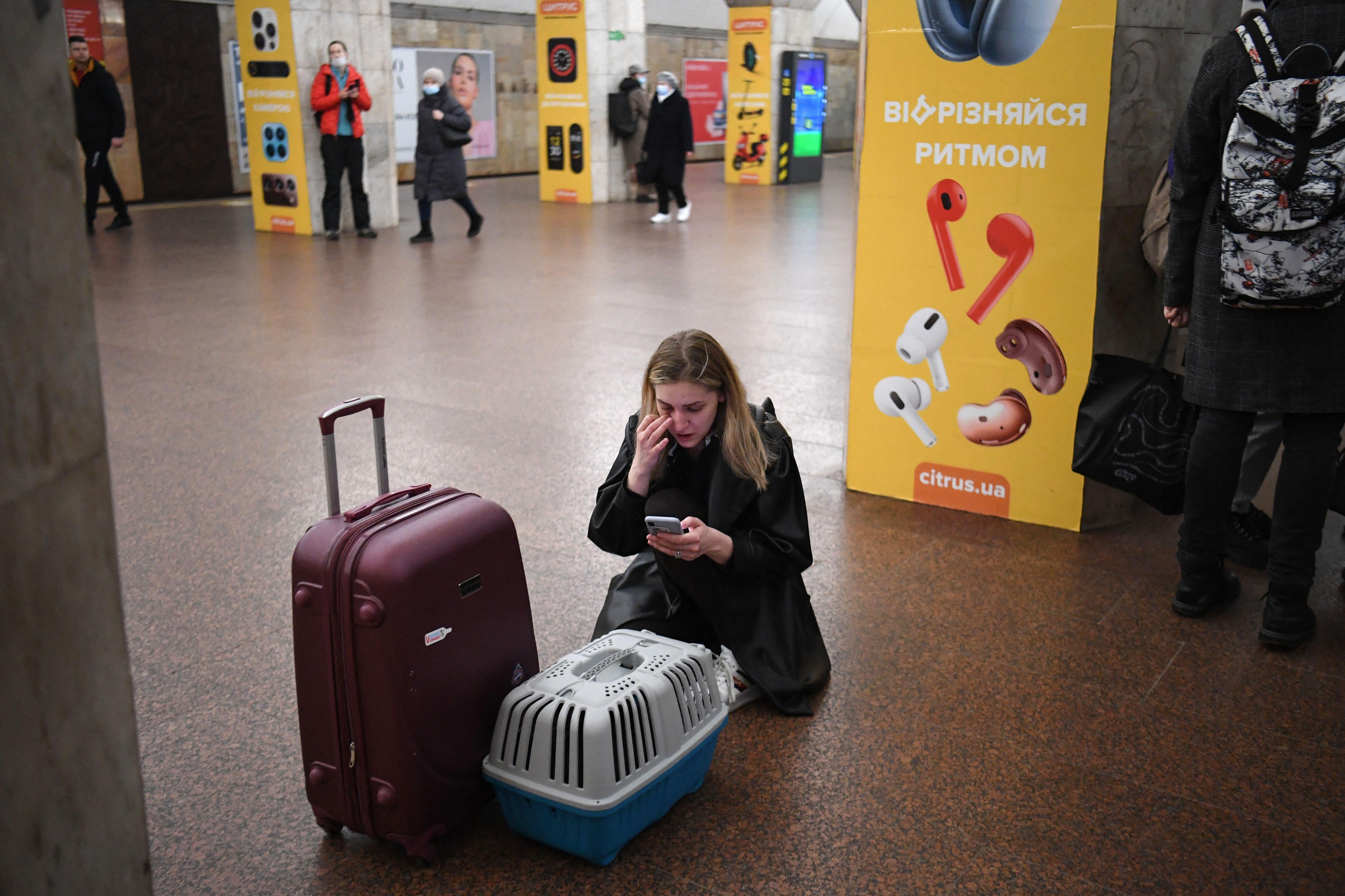 A woman in a subway station kneels on the floor in front of a suitcase and a cat carrier as she looks at her phone.