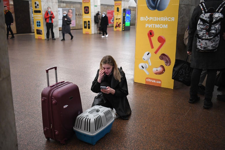 A woman in a subway station kneels on the floor in front of a suitcase and a cat carrier as she looks at her phone.