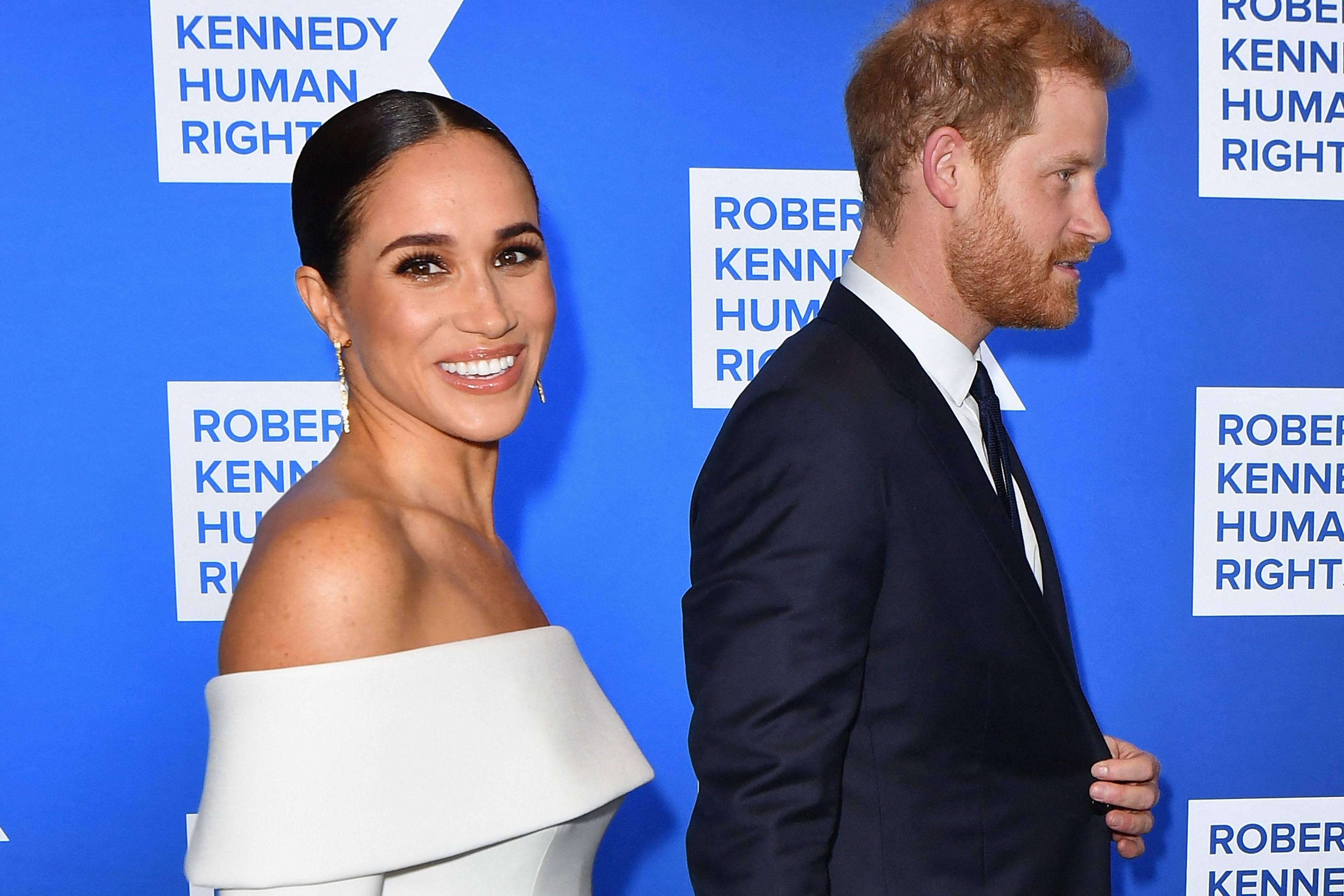 Harry and Meghan in front of a blue wall with logos for the Robert F. Kennedy Human Rights nonprofit.