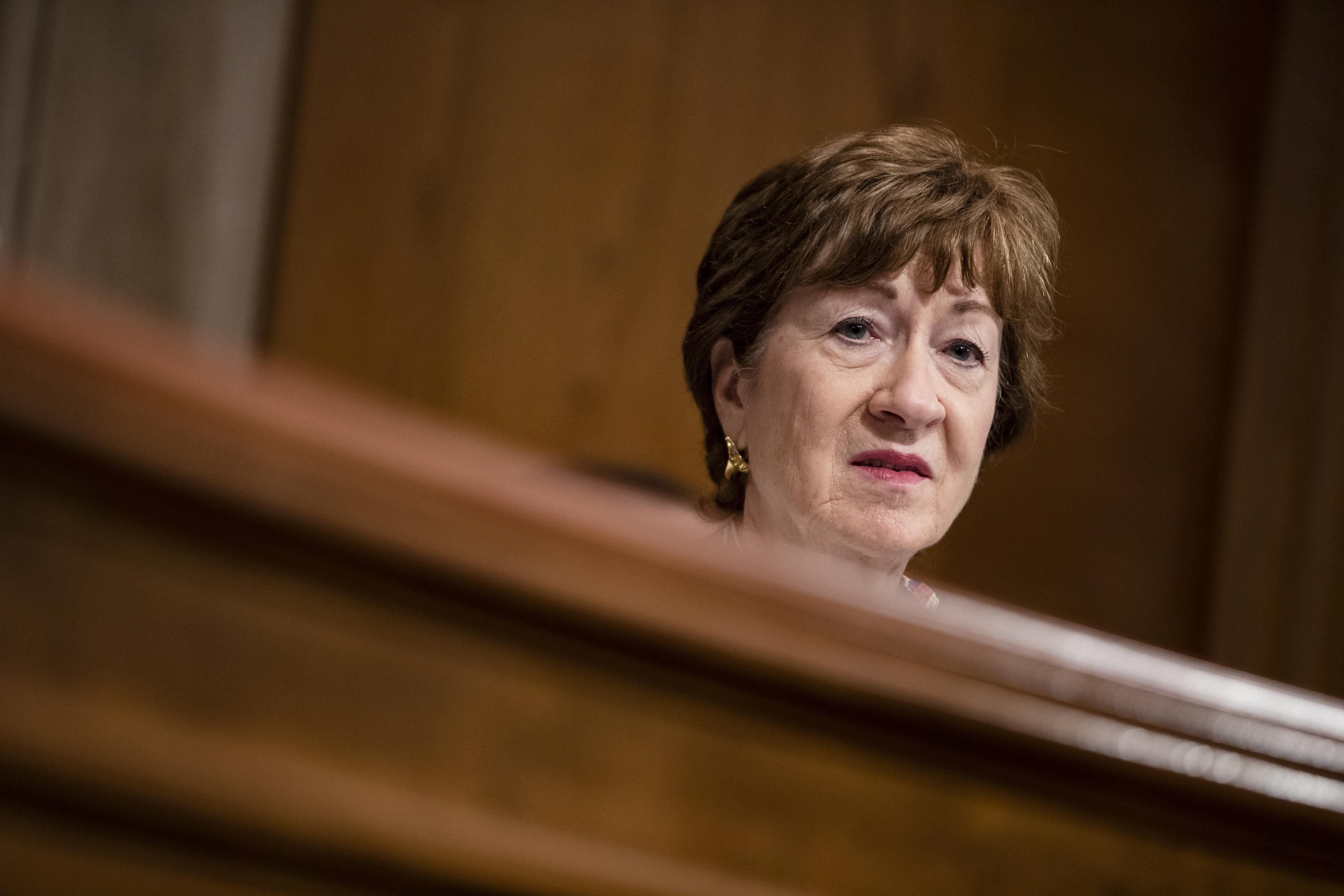 Susan Collins' head, wearing a solemn expression, peeks out above a wooden dais.
