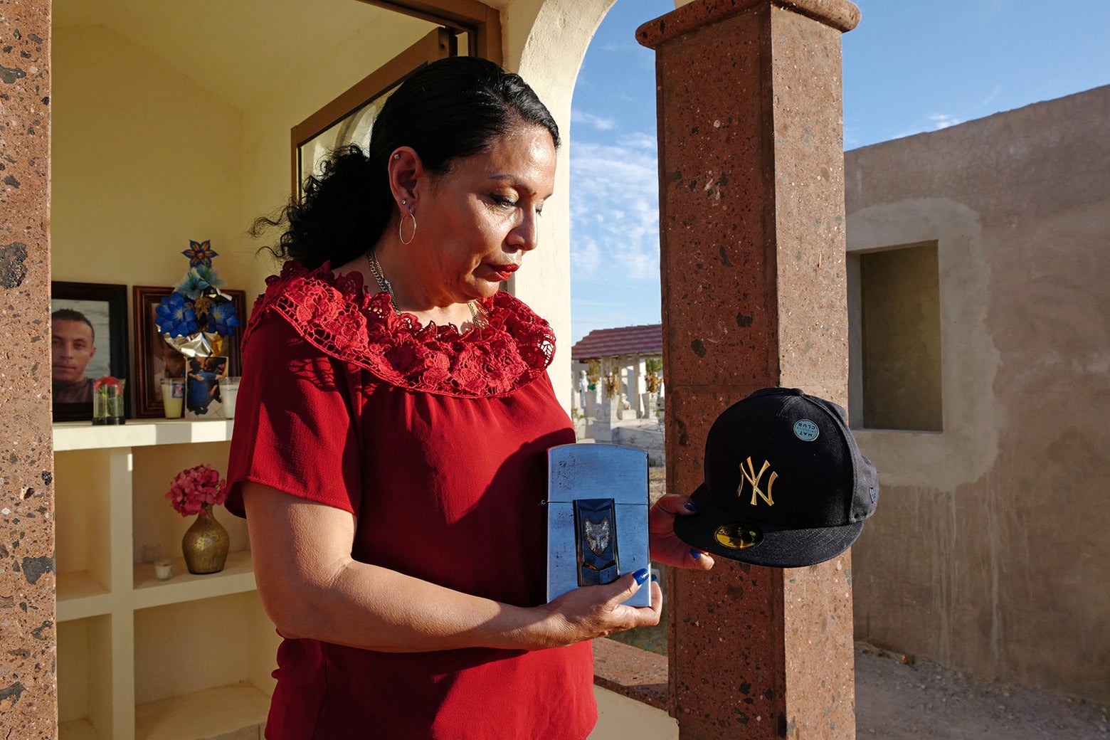 Cecilia, wearing a red dress, stands at her son's tomb in Hermosillo, Sonora, Mexico in May 2022 tearfullly holding a Yankees hat.