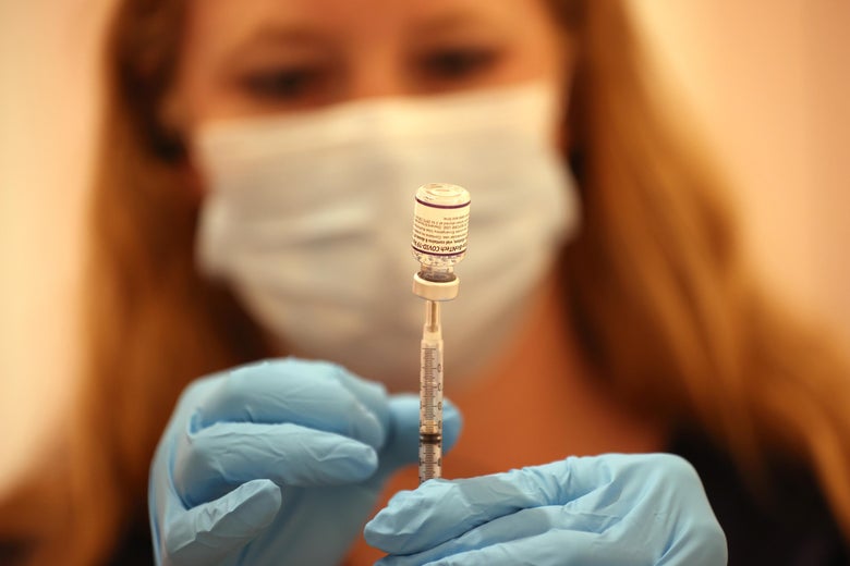 A woman in a surgical mask and blue gloves holds a syringe stuck in a glass vial.