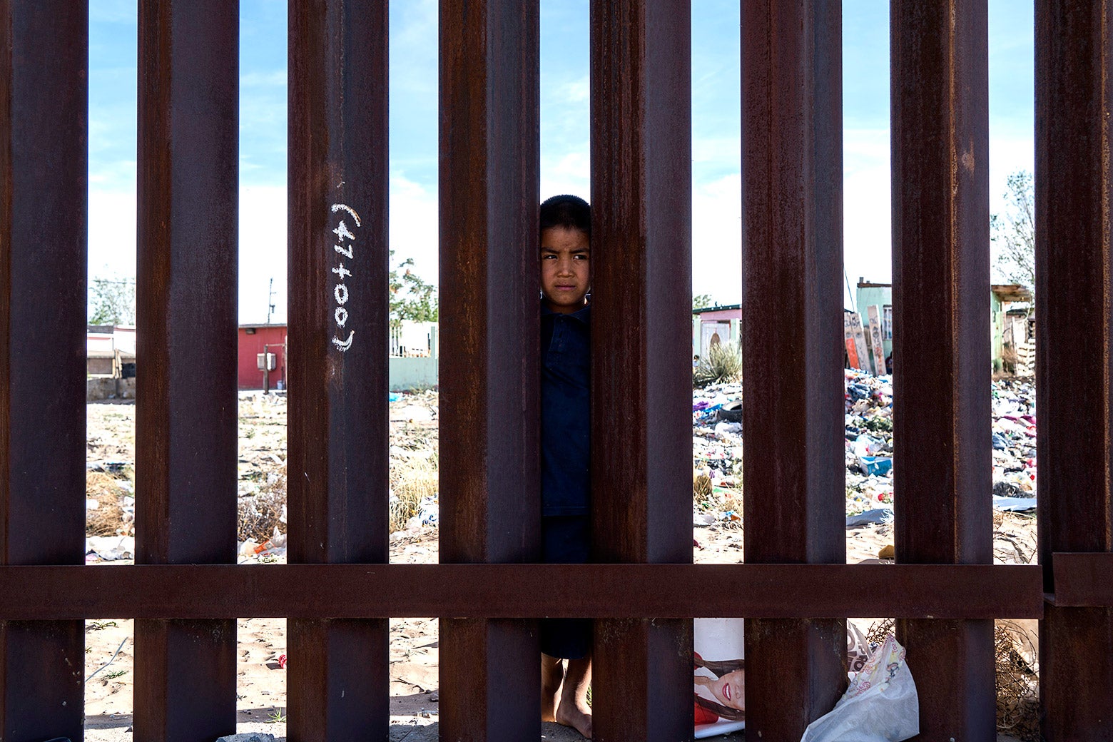 A young Mexican boy peers from the Mexican side of a border fence.
