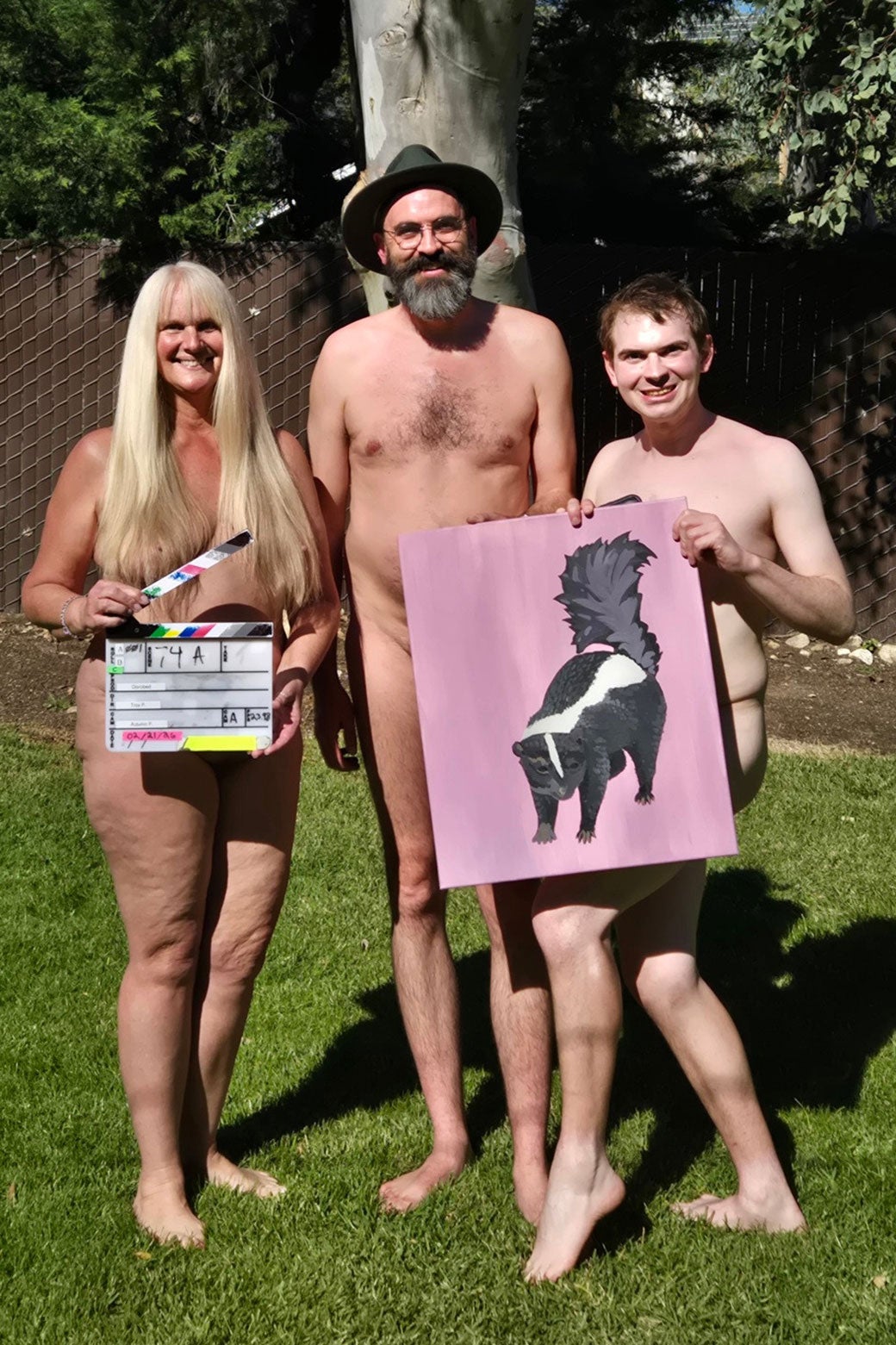 Three naked people with a director's clapperboard and a painting of a skunk hiding most of their bodies.