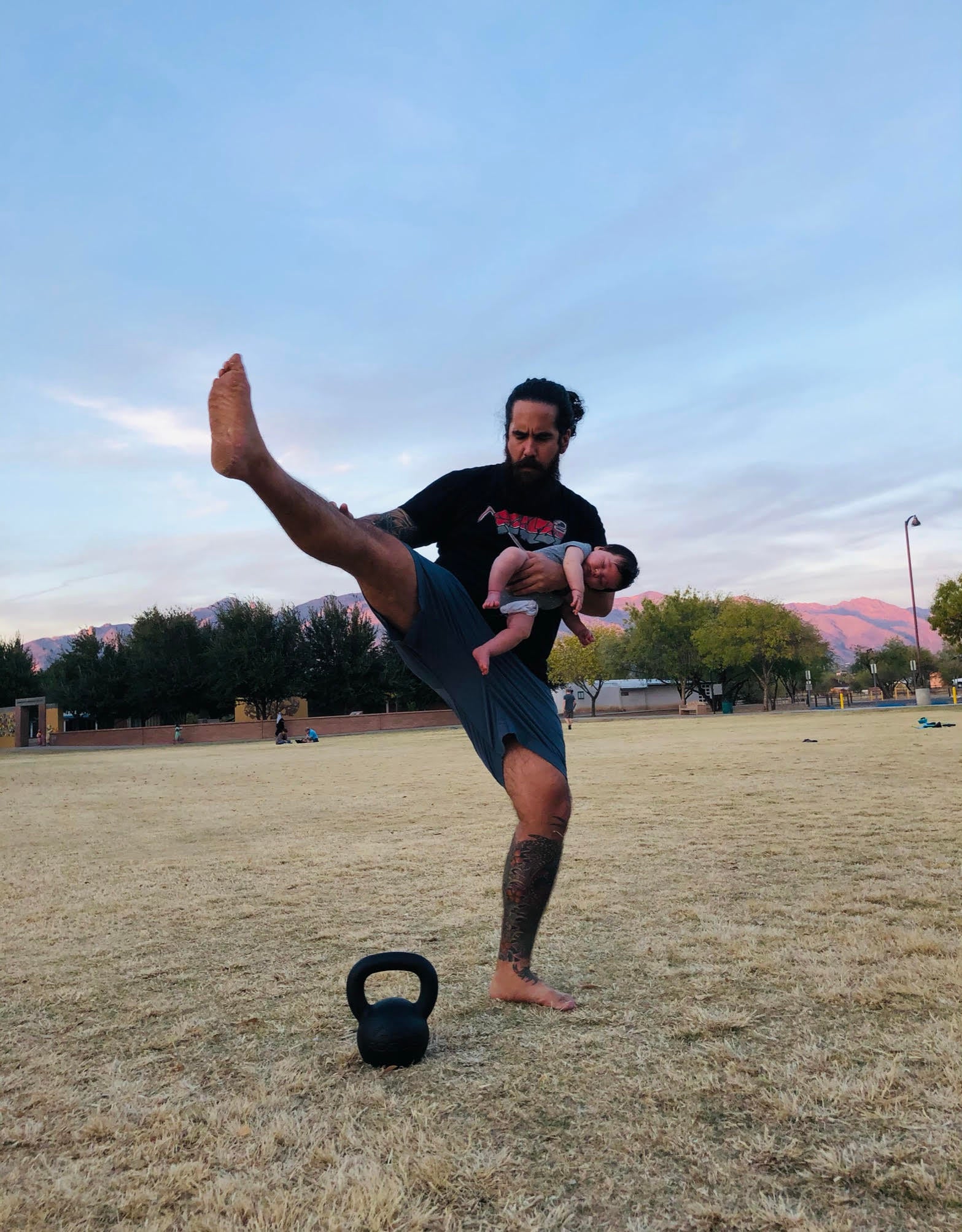 A man with a ponytail is exercising outside by kicking his right leg while holding a baby.