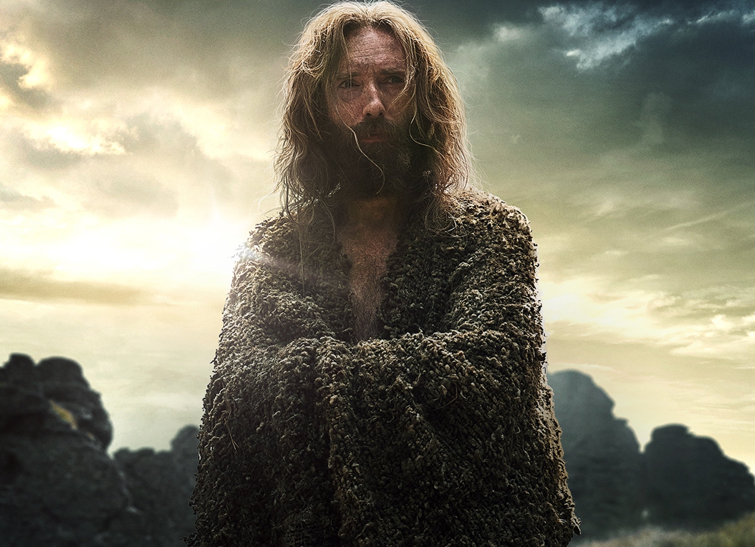 A long-haired man with a beard wrapped in a cloak stares into the distance.