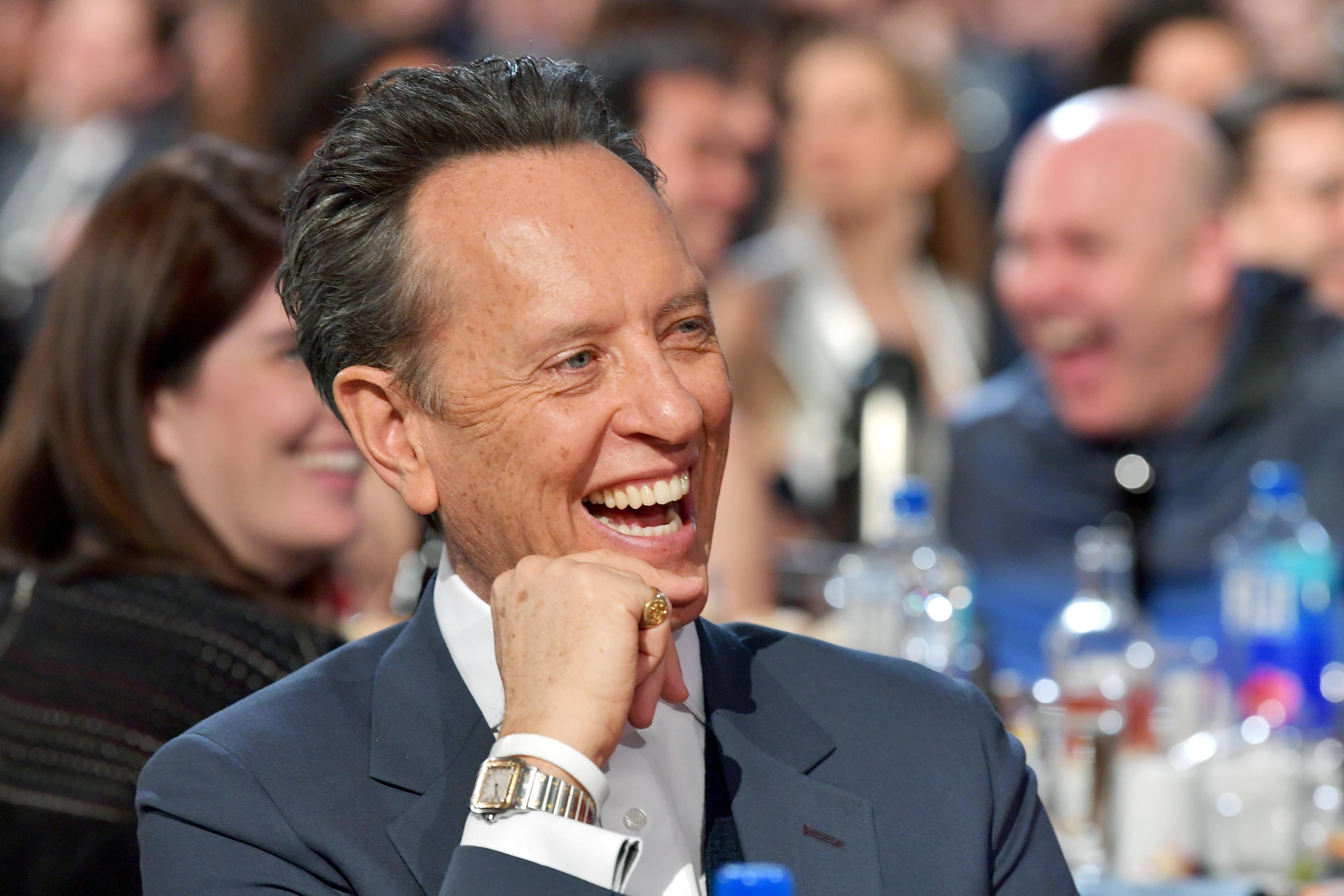 Richard E. Grant sitting at the Independent Spirit Awards, laughing.