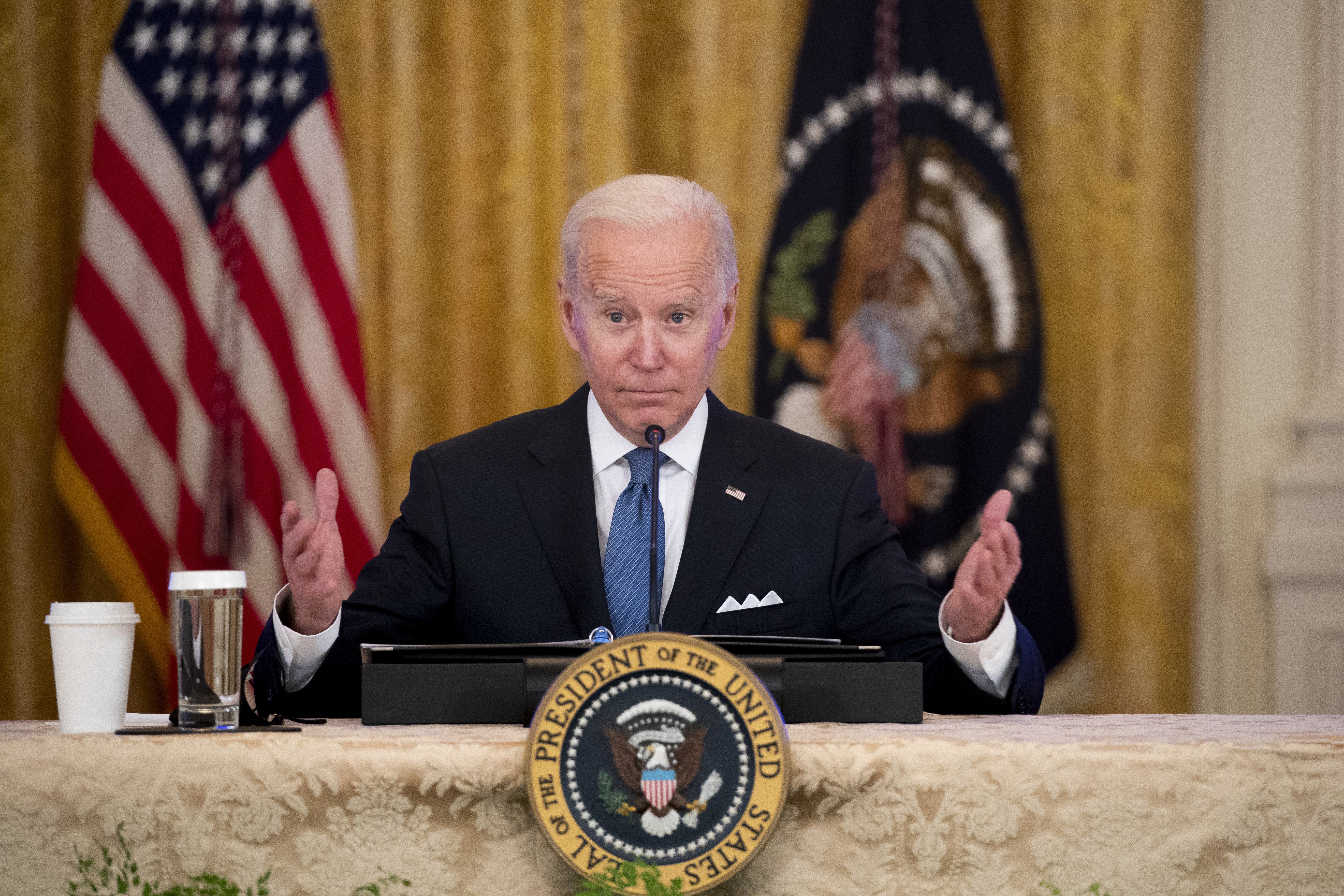 Biden gesturing with both hands as he sits with a presidential seal in front of him