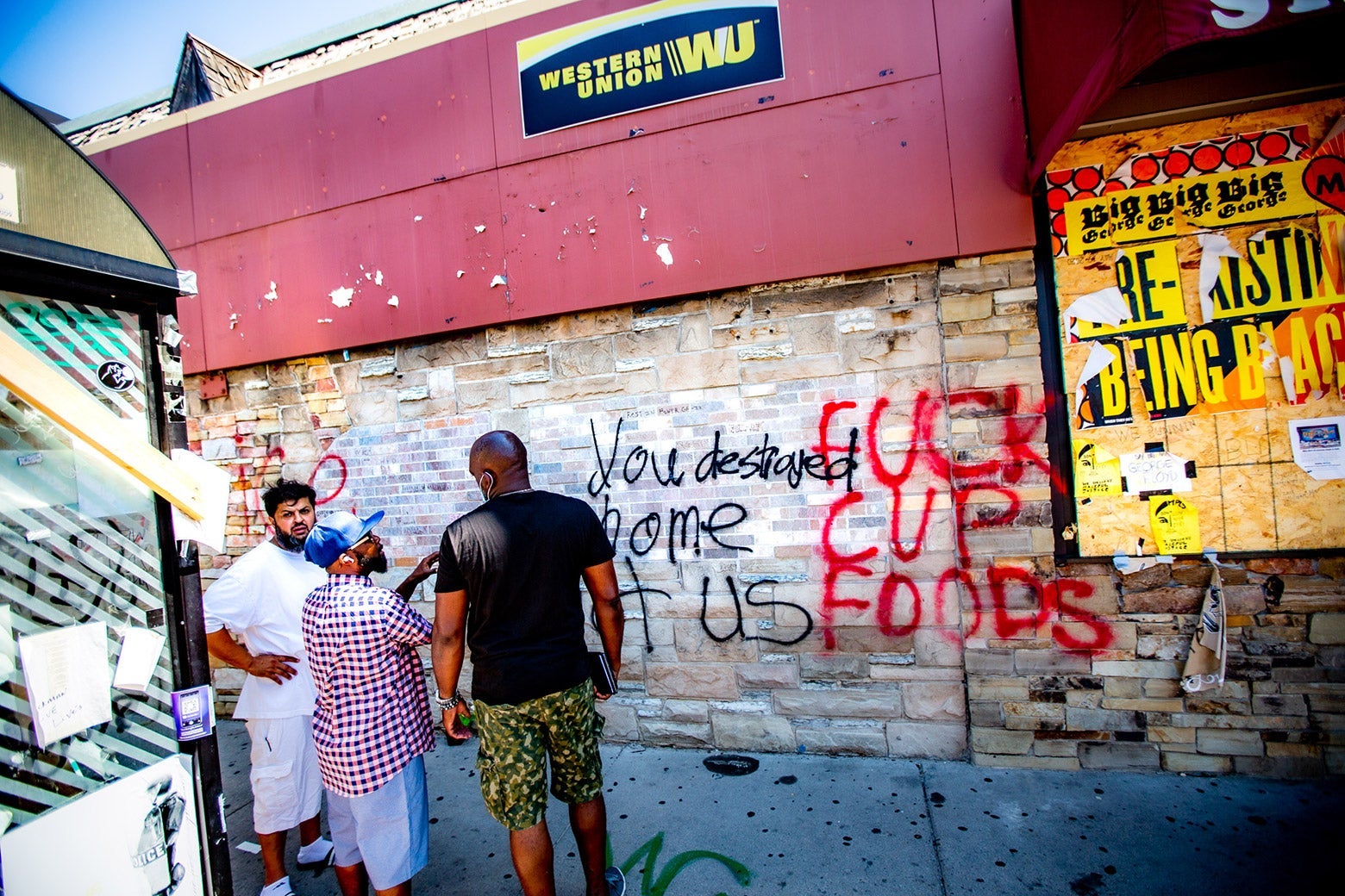 Three men stand in front of a wall covered in graffiti, saying things like, "FUCK CUP FOODS."