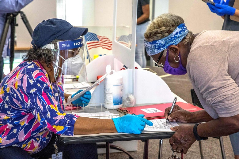 A poll worker, wearing a face mask, shield, and gloves, sits at a table and assists a mask-wearing voter as she hunches over to fill out her ballot.
