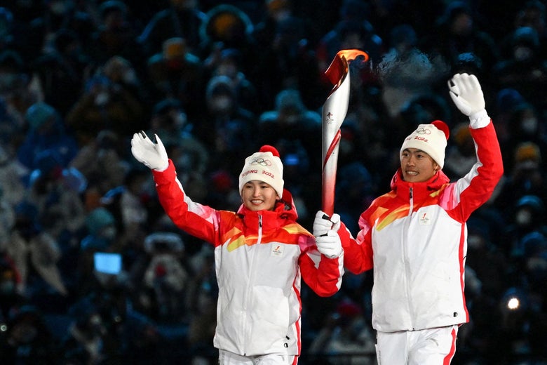 The athletes hold the torch together and smile, waving to the crowd