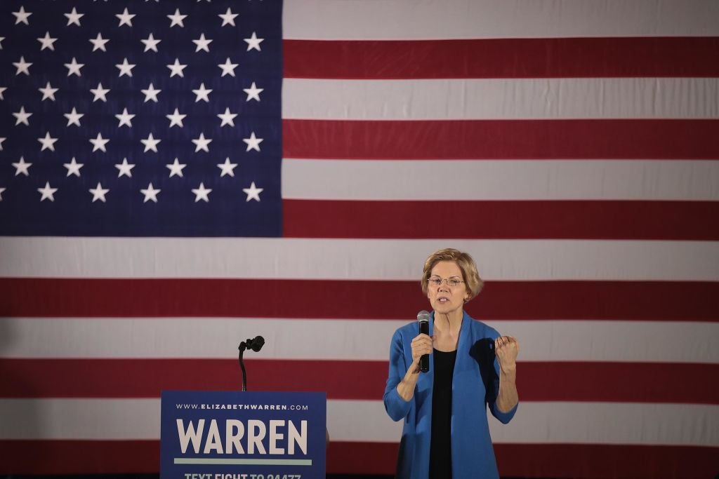Warren, wearing blue, speaks in front of a blue lectern with a WARREN sign on it and a large American flag backdrop.